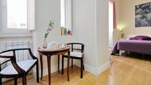 A room with a wooden floor, a bed with a purple bedspread, a chair, a small round table with a vase, and a window with a view of a building. - Amaranto Romano (Rome)
