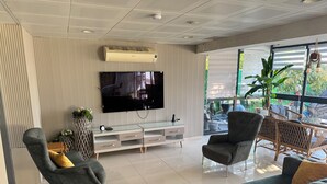 Ein modernes Wohnzimmer mit einem Flachbildfernseher, grauen Sofas, einem Glastisch und einem großen Fenster mit Blick auf Grünflächen.