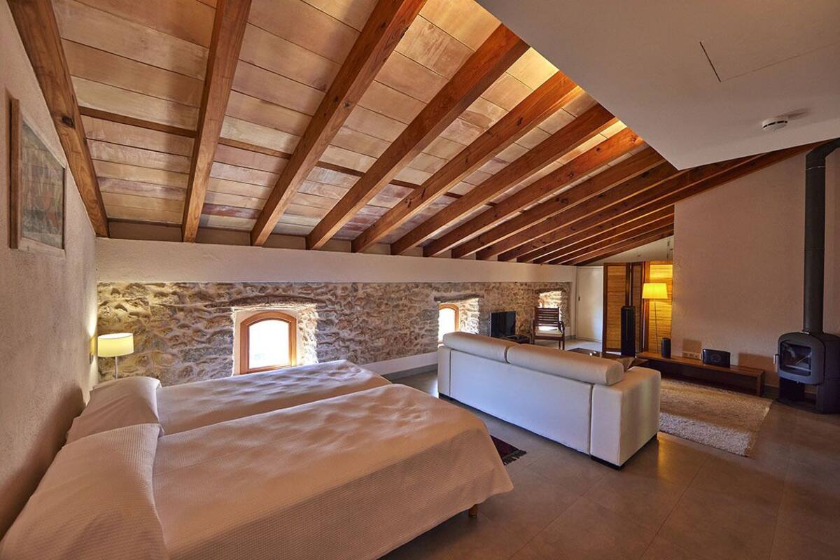 A bedroom with a stone wall, wooden beams, a sofa, and a fireplace.