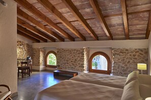 Un dormitorio con pared de piedra, vigas de madera, dos camas, un banco y un escritorio con lámpara.