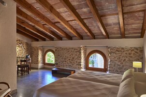 Un dormitorio con pared de piedra, vigas de madera, dos camas, un banco y un escritorio con lámpara.
