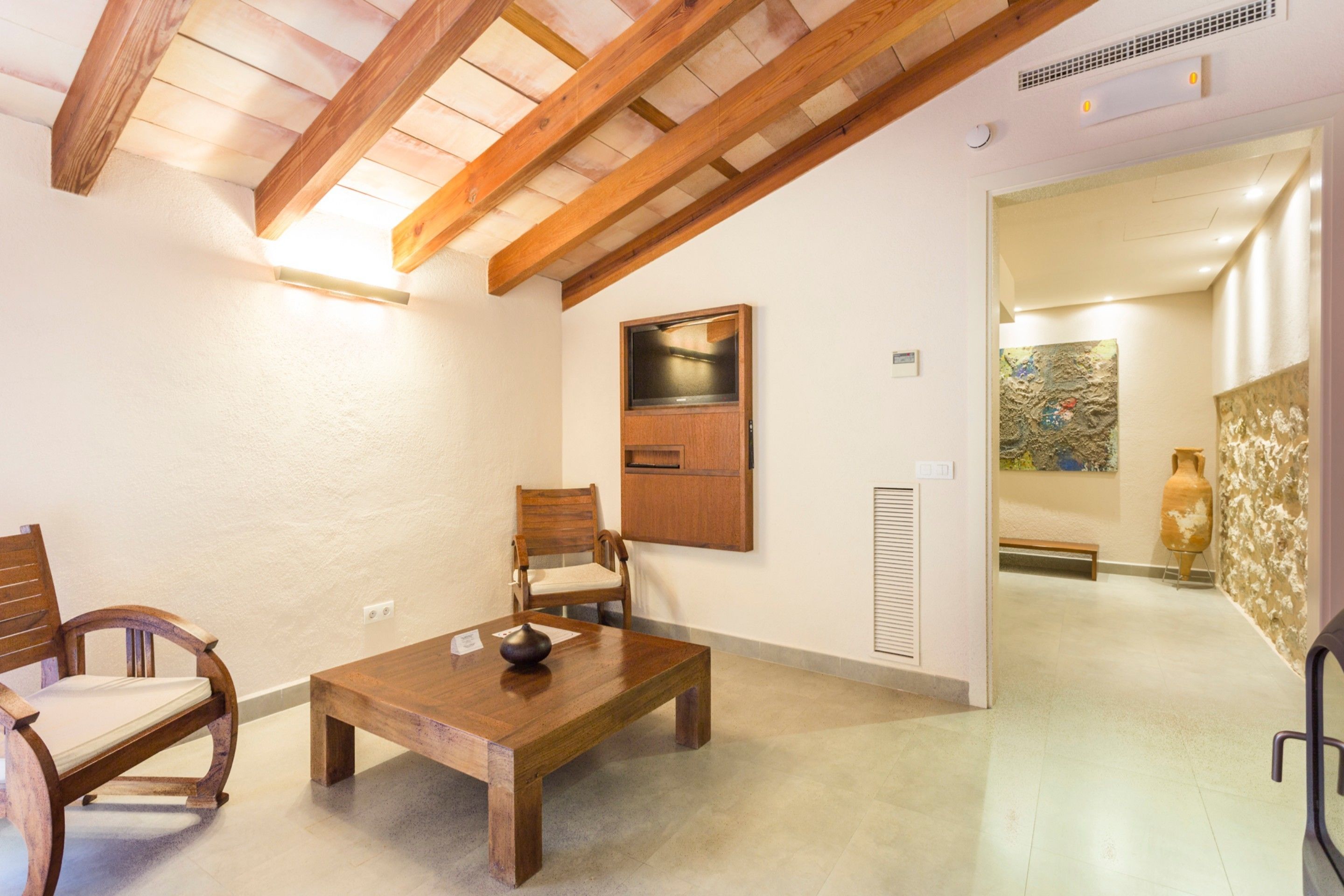 Quarto com mesa e cadeiras de madeira, teto de madeira, televisão presa na parede e um vaso decorativo.