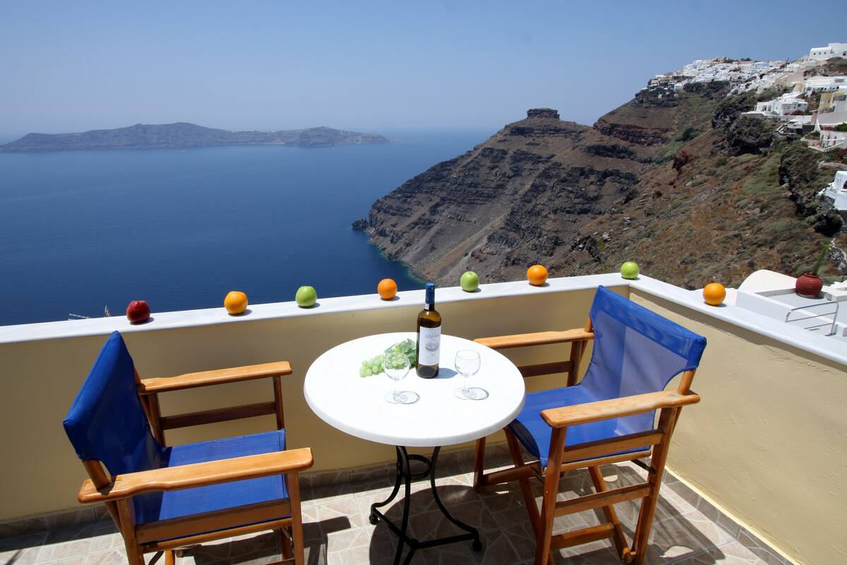 A balcony with two wooden chairs and a small table, overlooking a coastal view with a clear sky and distant islands.