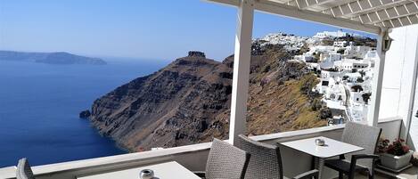A terrace with white tables and wicker chairs overlooking a coastal view with white buildings.