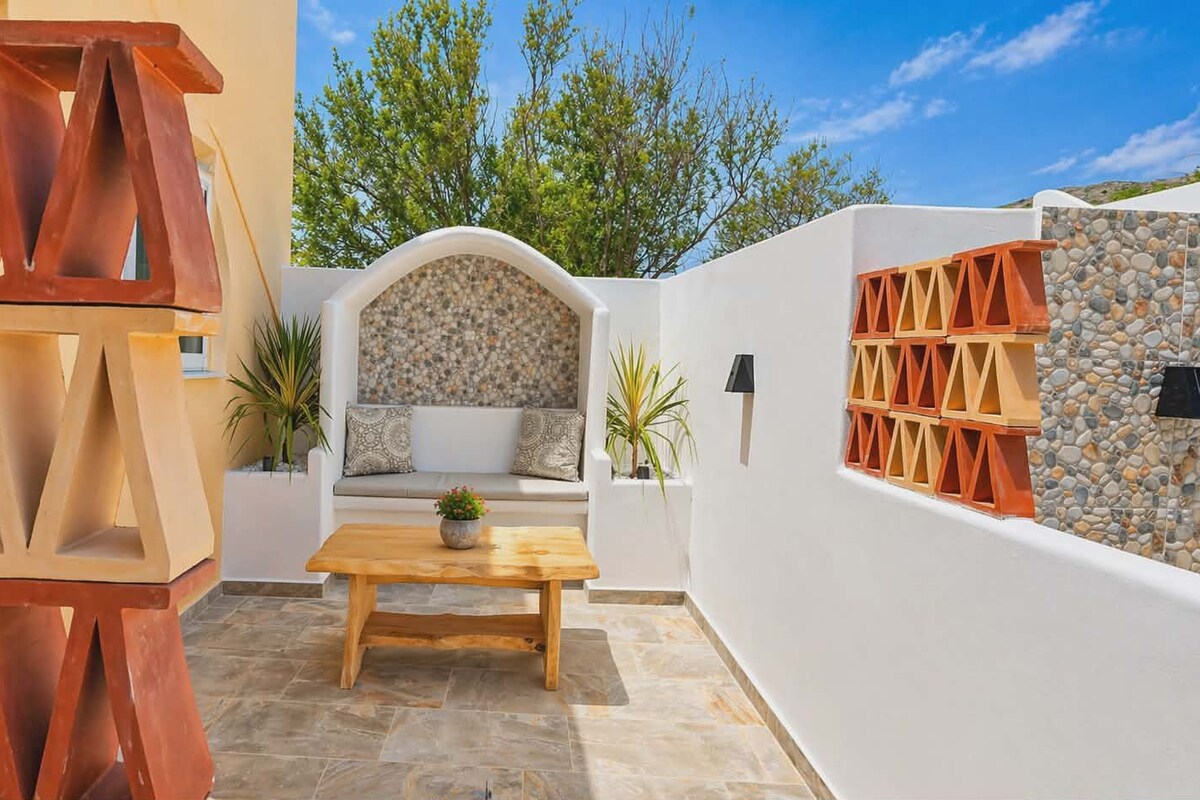 Un patio con una mesa de madera, un muro blanco con diseño de piedra y una zona de estar con cojines.