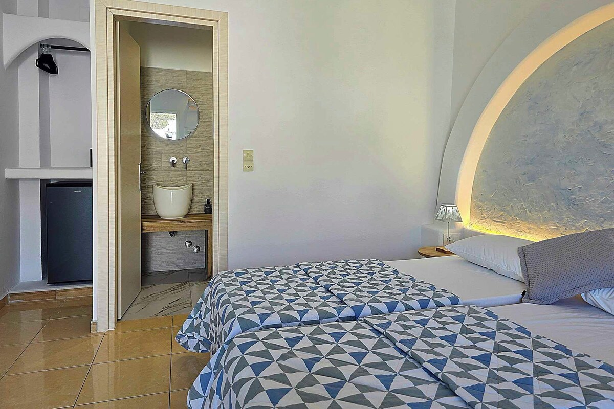 Un dormitorio con dos camas, un baño con lavamanos y espejo, y un refrigerador pequeño.