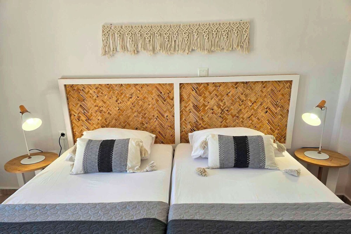 Habitación de hotel con dos camas, cada una con cabecero de madera, ropa de cama blanca y un tapiz de macramé en la pared. Hay dos mesitas de noche con lámparas.