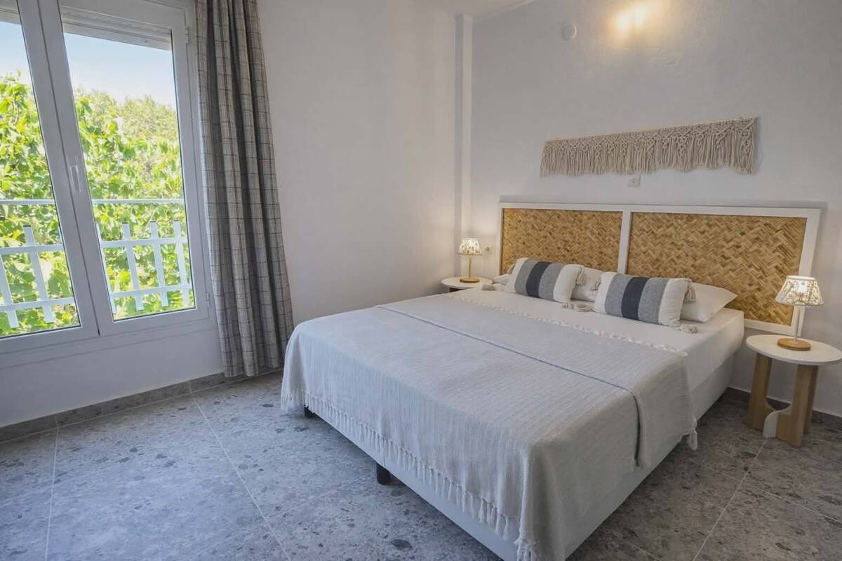 A bedroom with a large bed, a window with curtains, a wall-mounted macrame piece, and a small bedside table with a lamp.