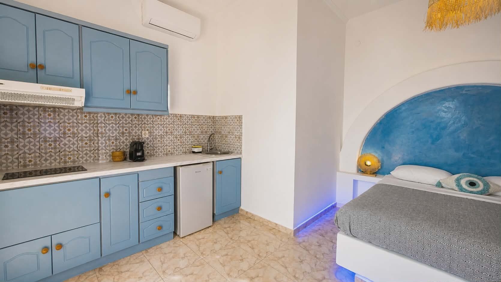 Una habitación compacta con kitchenette, una cama con cabecera azul y una mesita de noche pequeña.