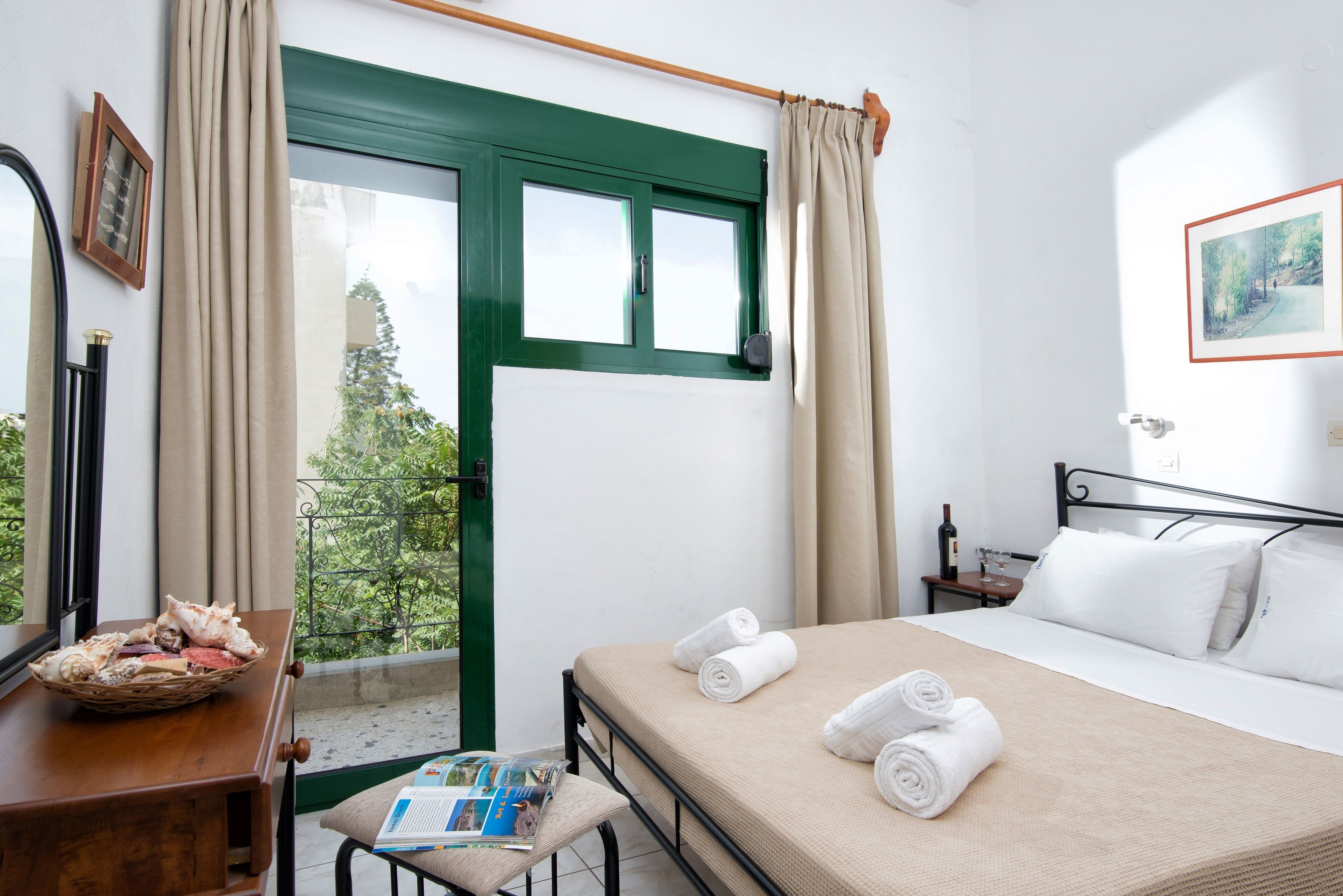 A hotel room with a bed, a window with curtains, a bedside table with a basket, a magazine, and a framed picture on the wall.