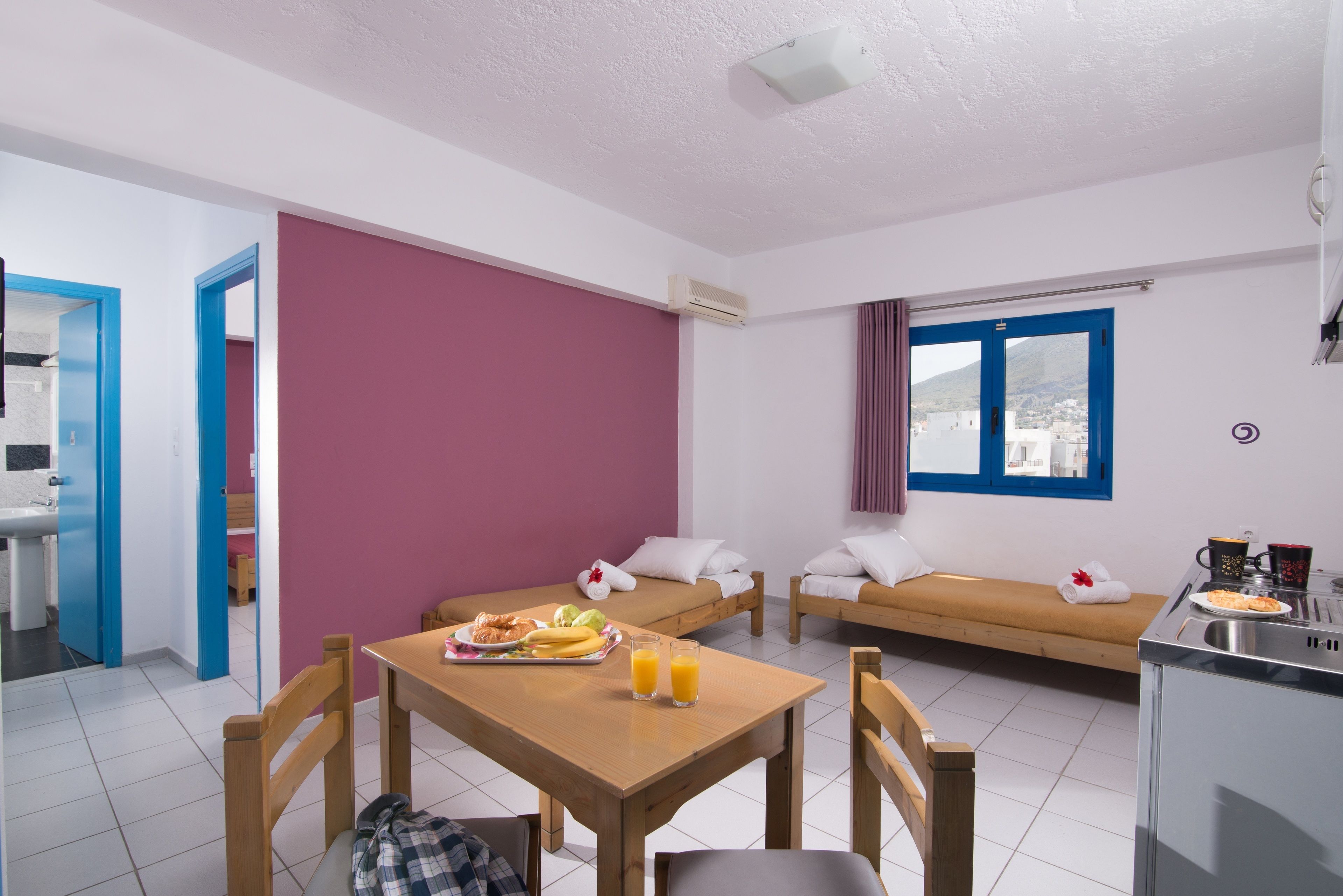 Une pièce compacte comprenant une table à manger, deux lits avec des oreillers, une fenêtre avec des rideaux et un coin cuisine.