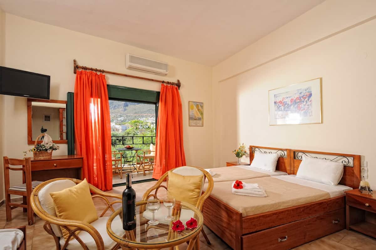 A hotel room with a bed, a desk with a chair, a small table with a bottle and glasses, and a balcony with orange curtains.