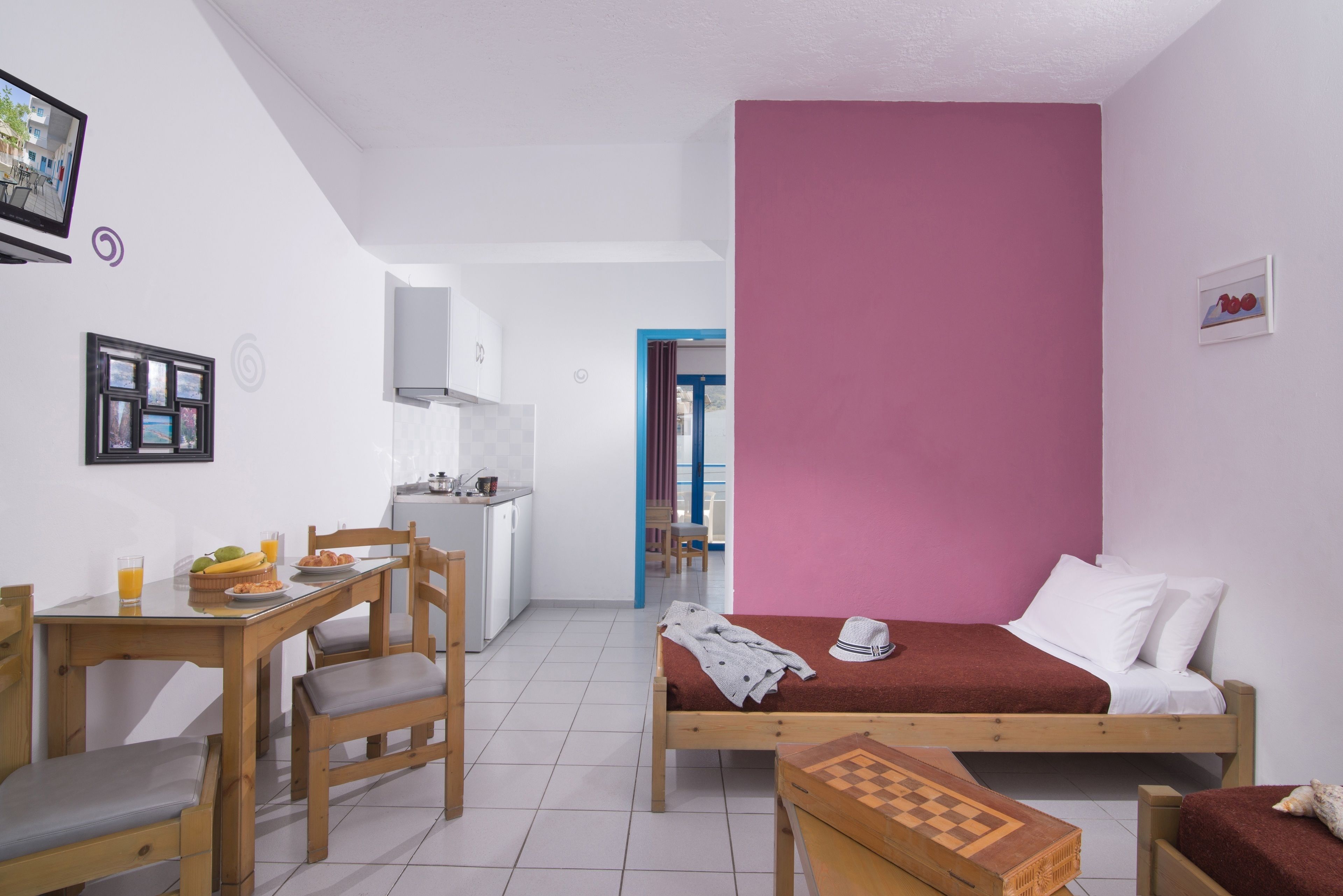 Ein kompakter Raum mit Bett, kleinem Schreibtisch, Stuhl und Esstisch. Es gibt eine Küchenzeile mit Schränken und Spüle.