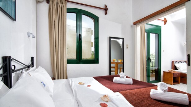 A hotel room with two beds, a large window with curtains, a mirror, a wooden door, and a small table with a chair. - Erofili Apartments (Hersonissos)