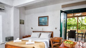 A bedroom with a bed, bedside tables, a wooden chair, a dining table with fruit and glasses, and a painting on the wall. - Erofili Apartments (Hersonissos)