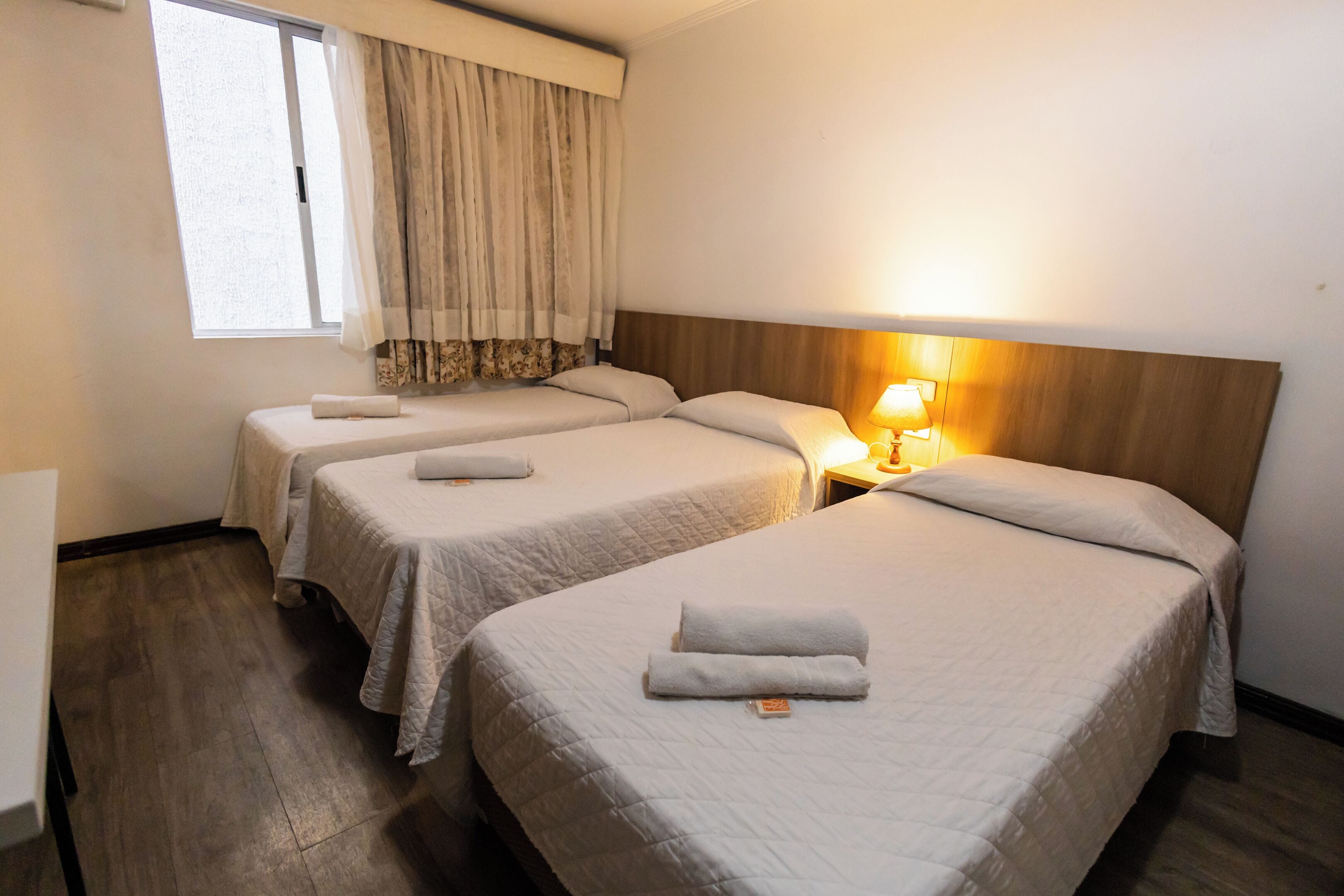 Habitación de hotel con dos camas individuales, una ventana con cortinas, una lámpara de noche y un cabecero de madera.