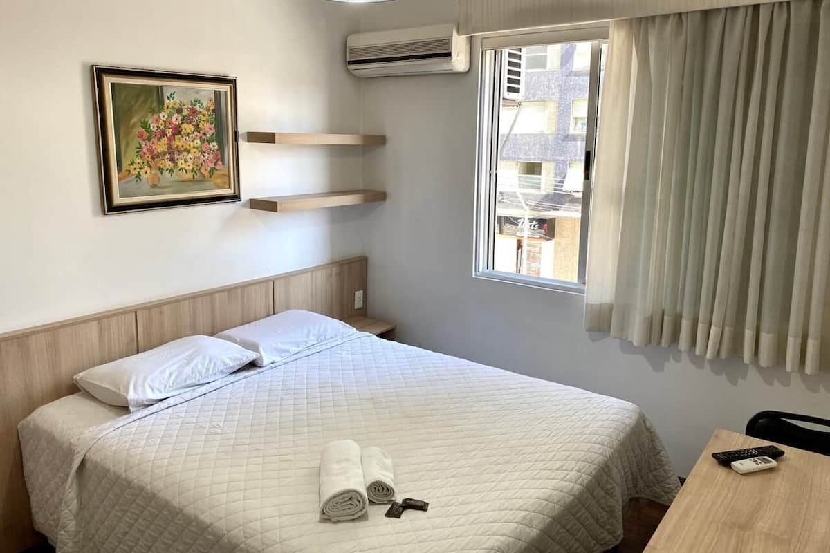 A hotel room with a bed, a painting on the wall, a window with curtains, and a bedside table with a remote control.