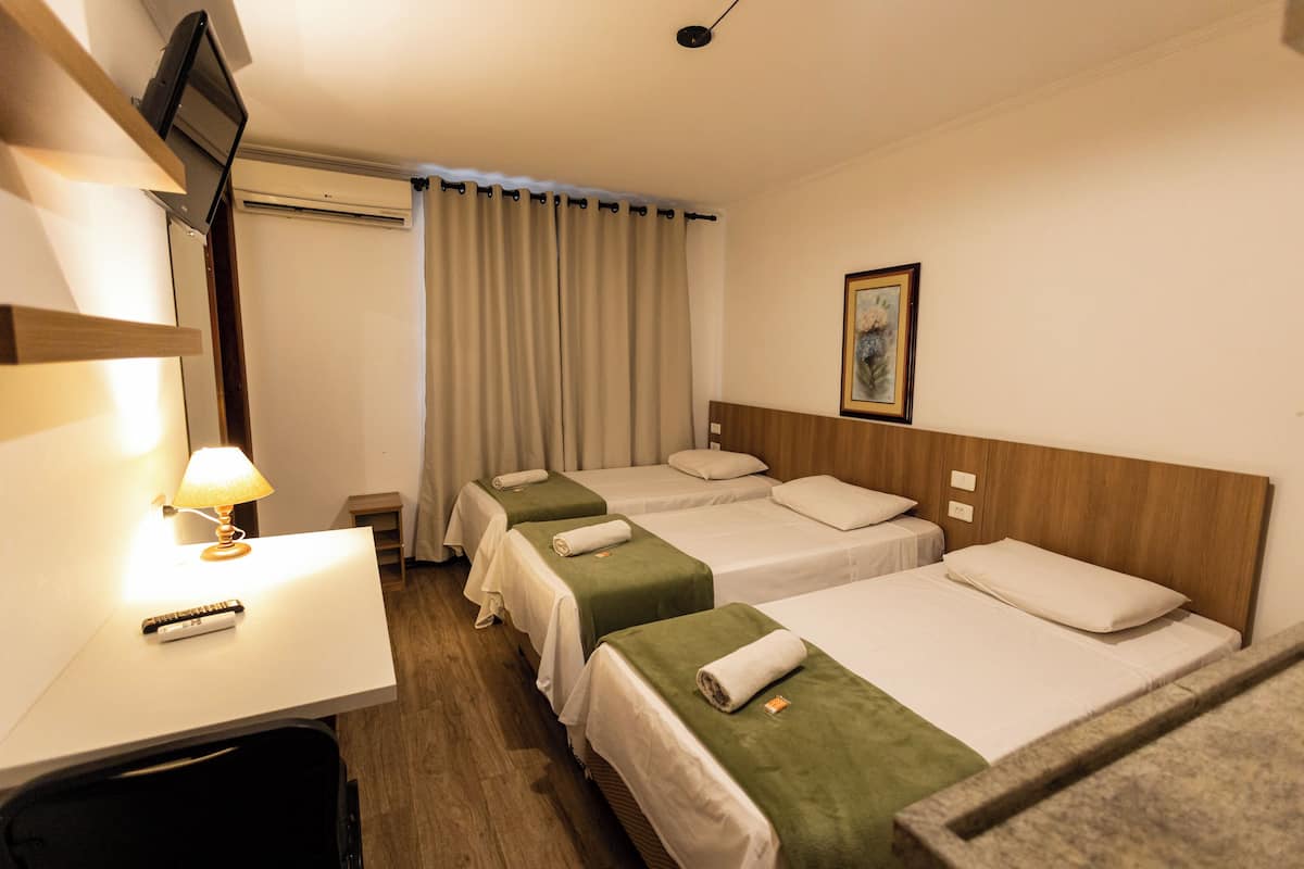 A hotel room with three beds, a desk with a lamp, a TV, and a painting on the wall.
