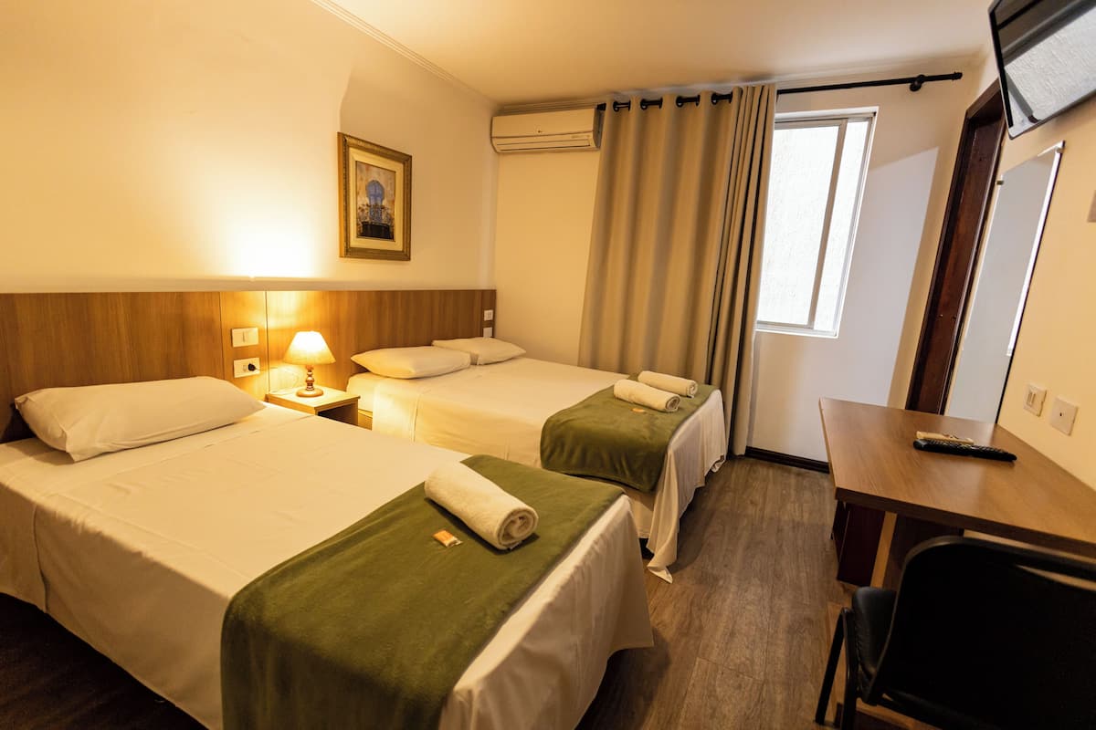 A hotel room with two beds, a desk, a chair, a lamp, an air conditioner, and a window with curtains.