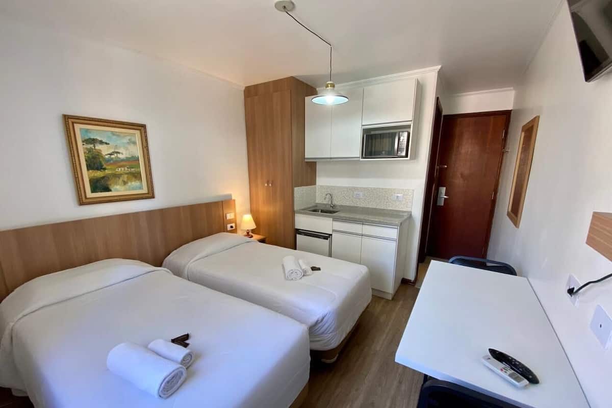 A hotel room with two beds, a kitchenette, a desk, and a painting on the wall.