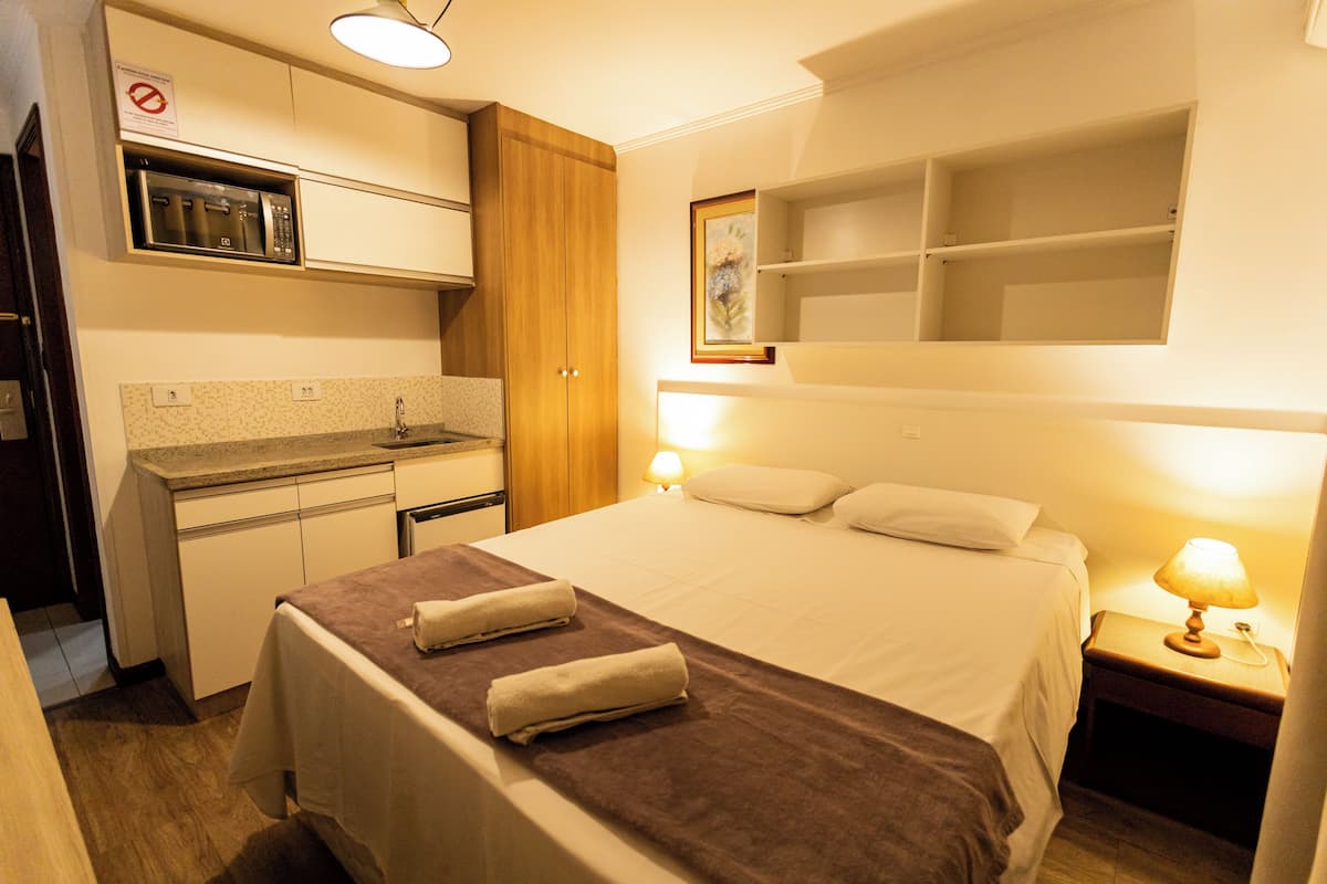 A hotel room with a bed, a nightstand with a lamp, a wooden wardrobe, a kitchenette with a microwave, and a framed picture on the wall.