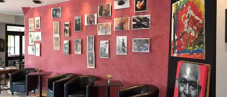 A lobby with a red wall adorned with framed photographs and a large abstract painting. There are black leather chairs and a small table with a vase.