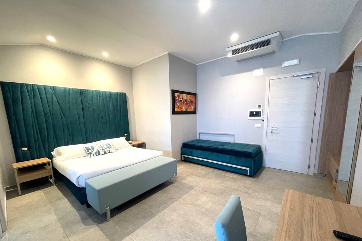 Ein modernes Hotelzimmer mit Bett, Nachttischen, einer Bank, einem Schreibtisch und einer Klimaanlage.