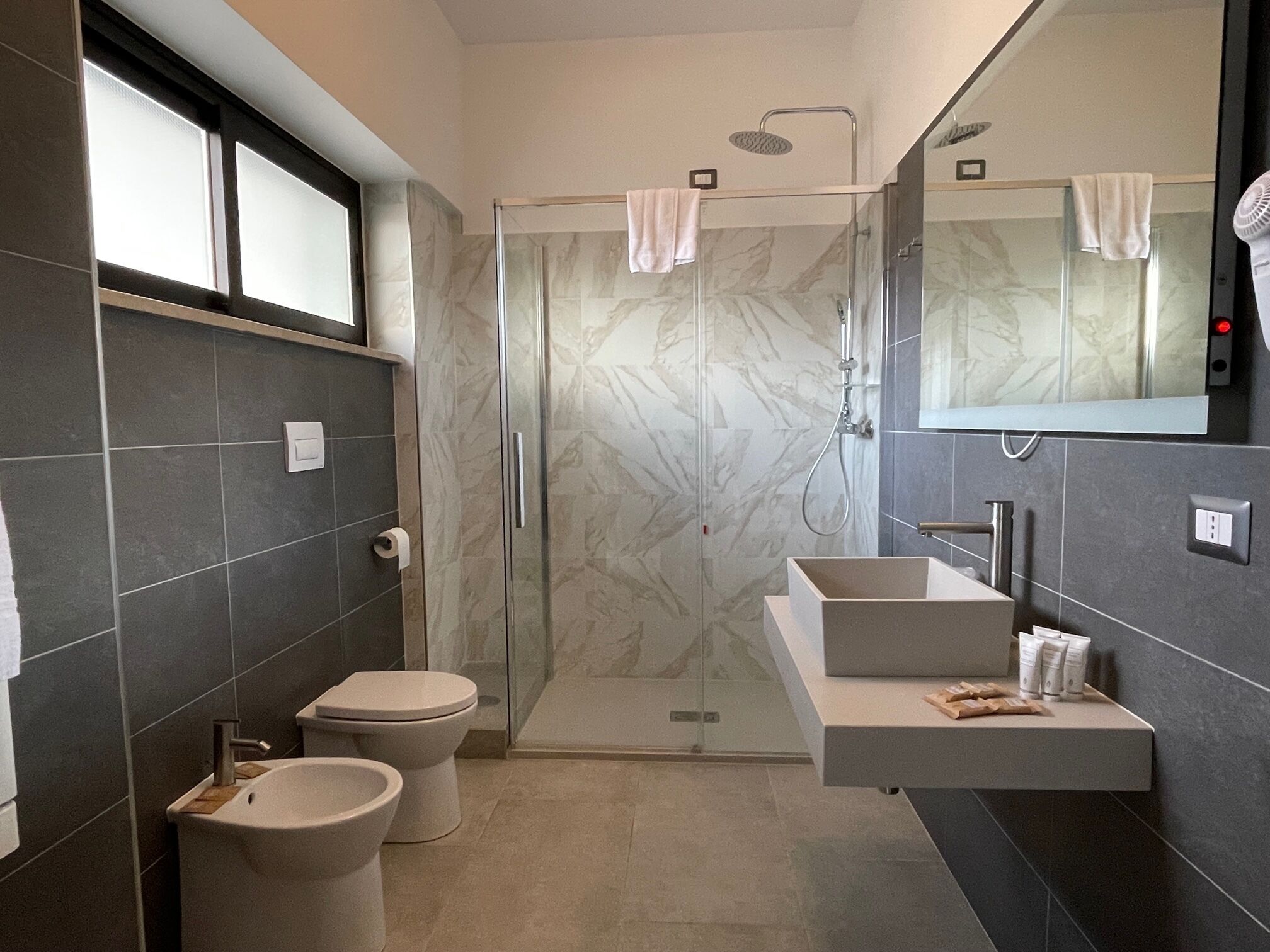 A modern bathroom with a glass-enclosed shower, a vanity with a large mirror, and two toilets.