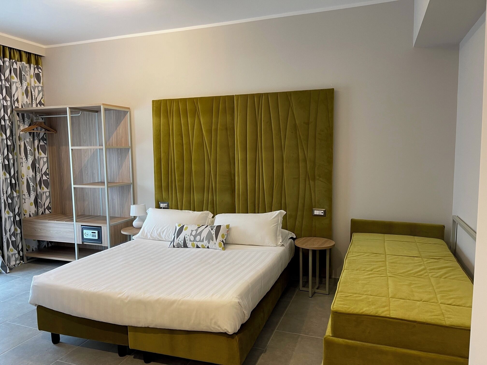 Een hotelkamer met een groot bed, een kleiner bed, een nachtkastje, een kledingkast en een kapstok.