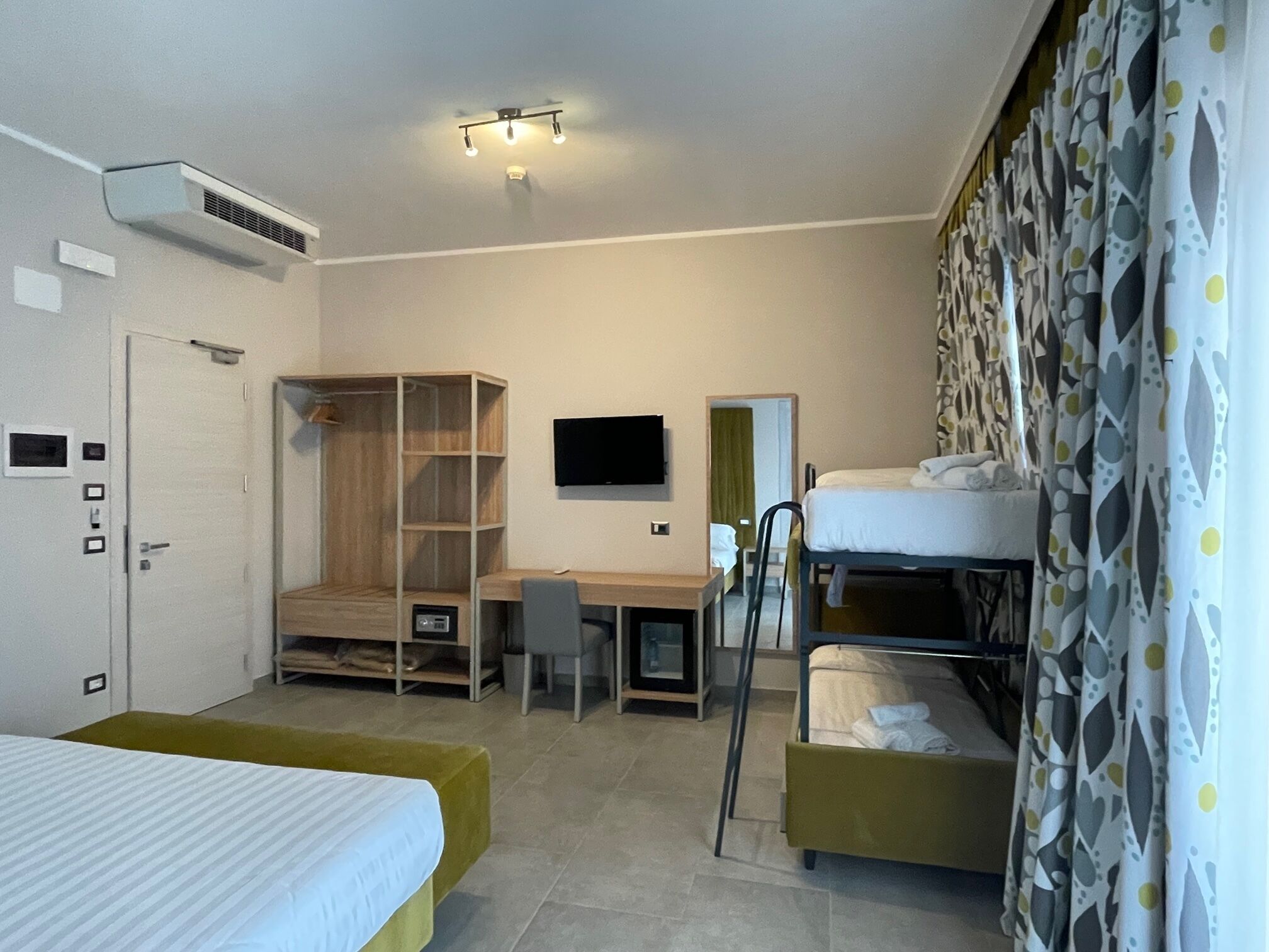 Una camera d'albergo con un letto a castello, una scrivania con sedia, una TV a parete e un armadio.