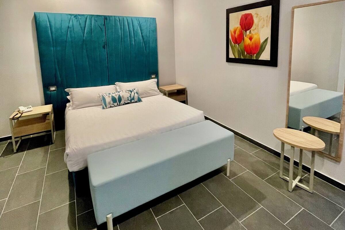 Ein modernes Schlafzimmer mit einem großen Bett, einem Nachttisch, einem Wandspiegel und einem gerahmten Bild von Blumen.