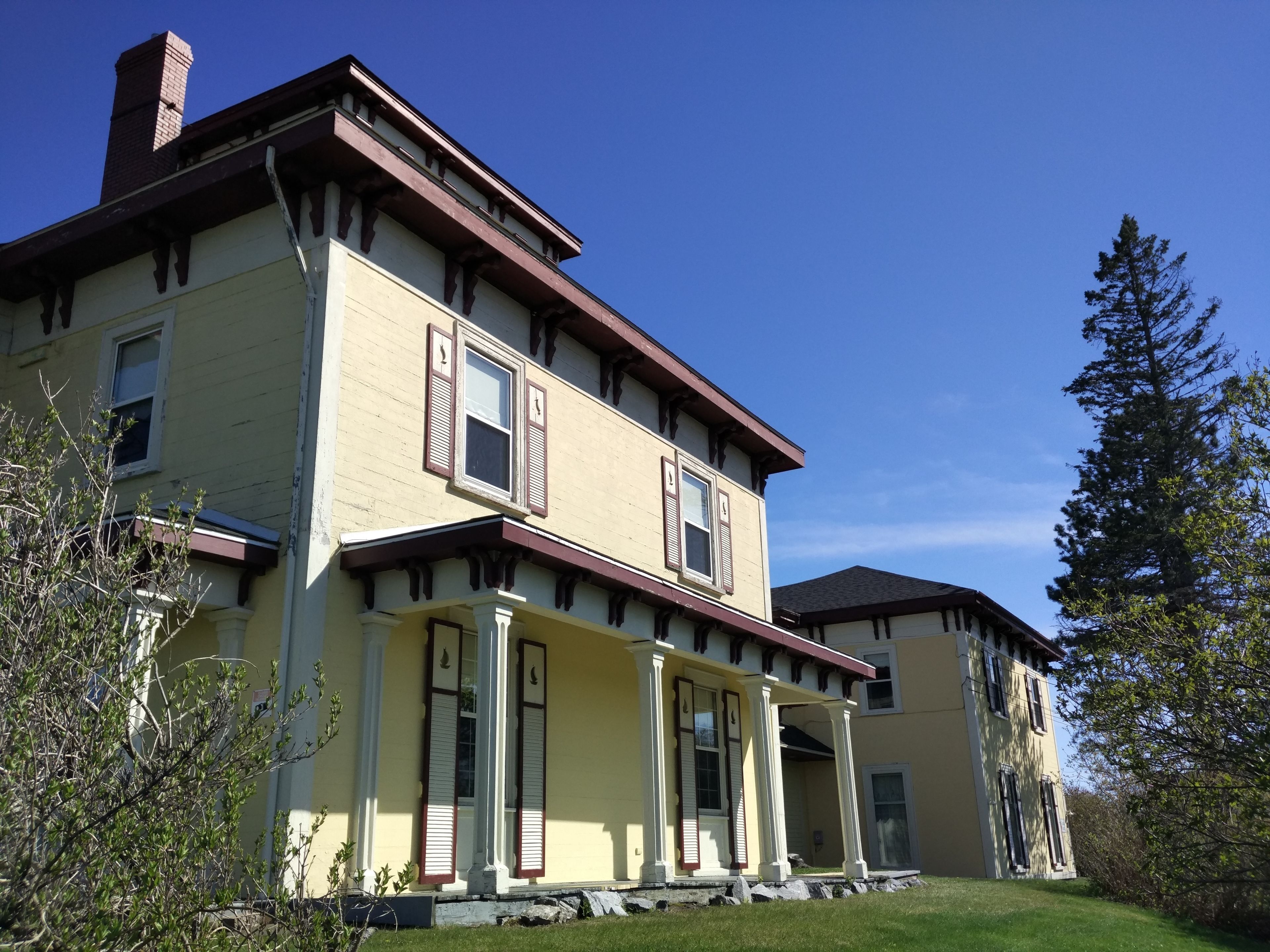A two-story yellow house with a porch, a chimney, and a large tree in the background.