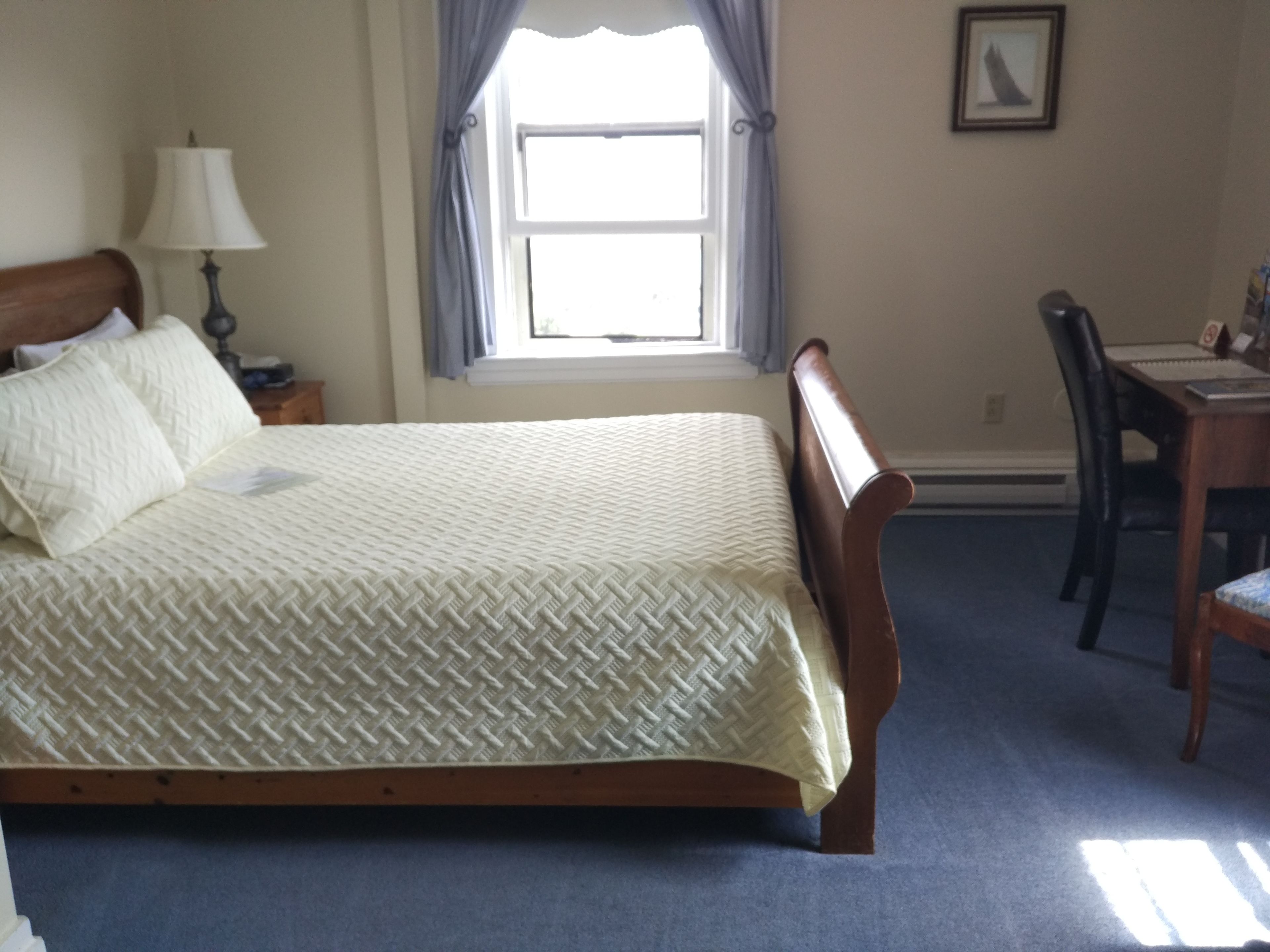 Une chambre à coucher avec un lit, une table de chevet avec une lampe, une fenêtre avec des rideaux, un bureau avec une chaise et un tableau encadré au mur.