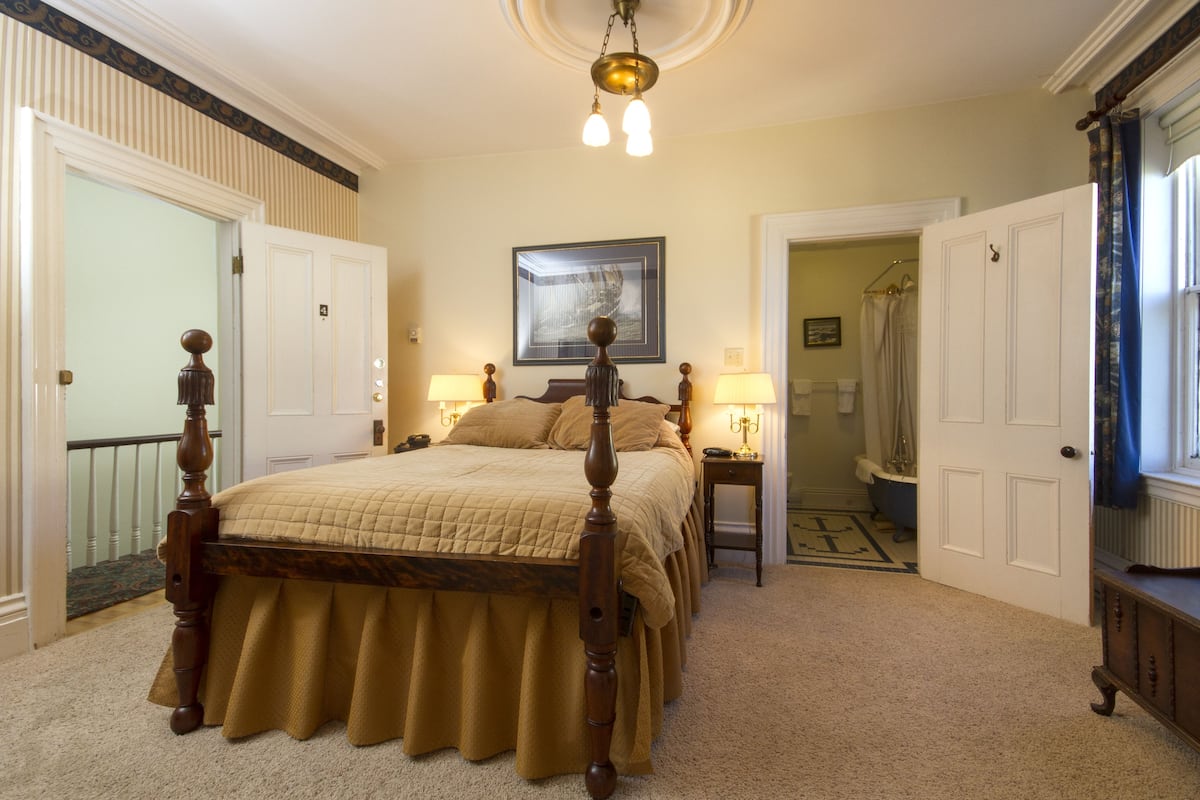 Une chambre à coucher avec un grand lit, une table de chevet, une lampe, un tableau encadré au mur et une porte donnant sur une autre pièce.