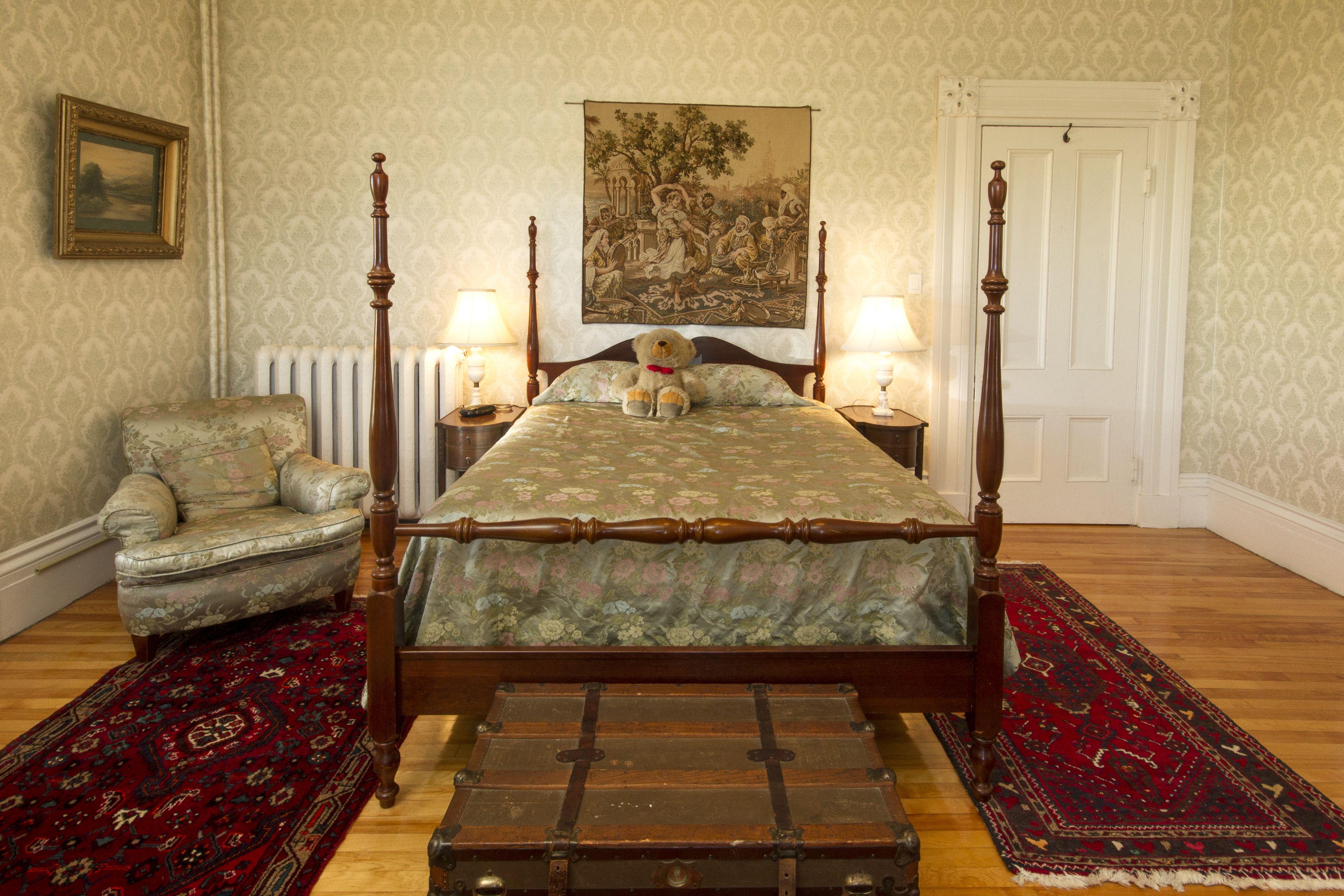 A bedroom with a four-poster bed, a floral armchair, a red and black patterned rug, and a vintage trunk.