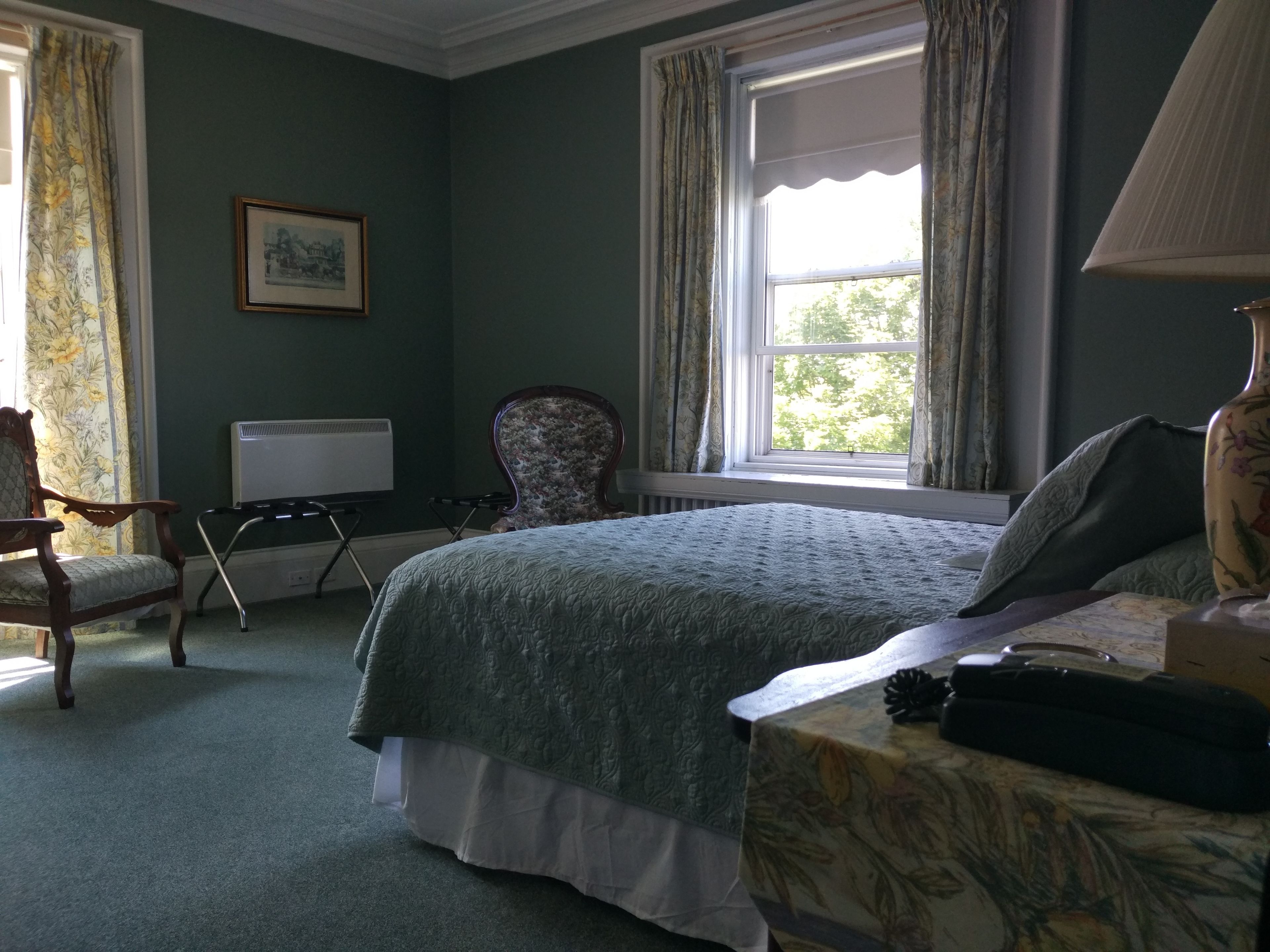 A bedroom with a bed, a chair, a radiator, a window with curtains, and a framed picture on the wall.