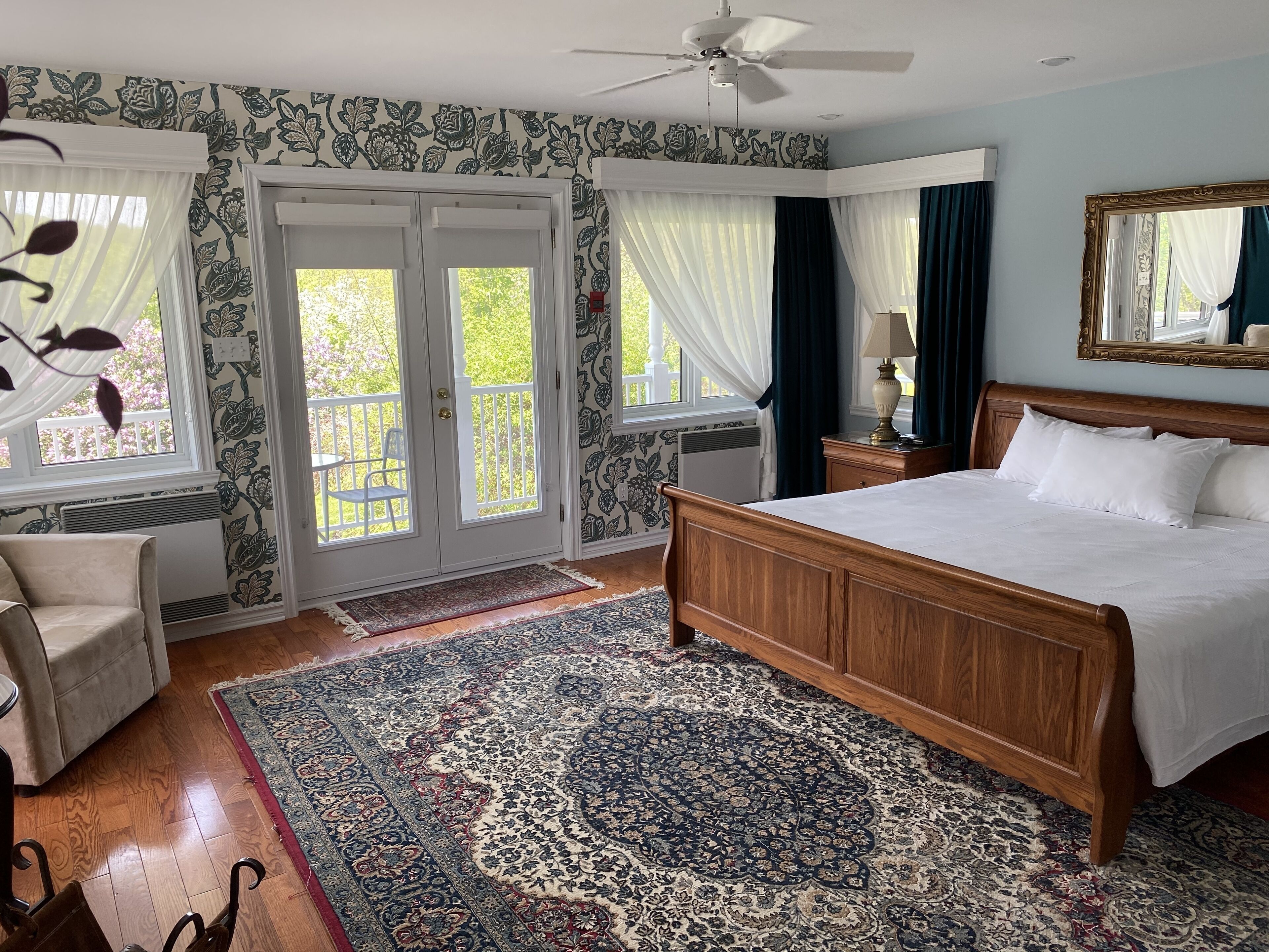 A bedroom with a large bed, a ceiling fan, a wooden headboard, a nightstand with a lamp, and a large mirror.