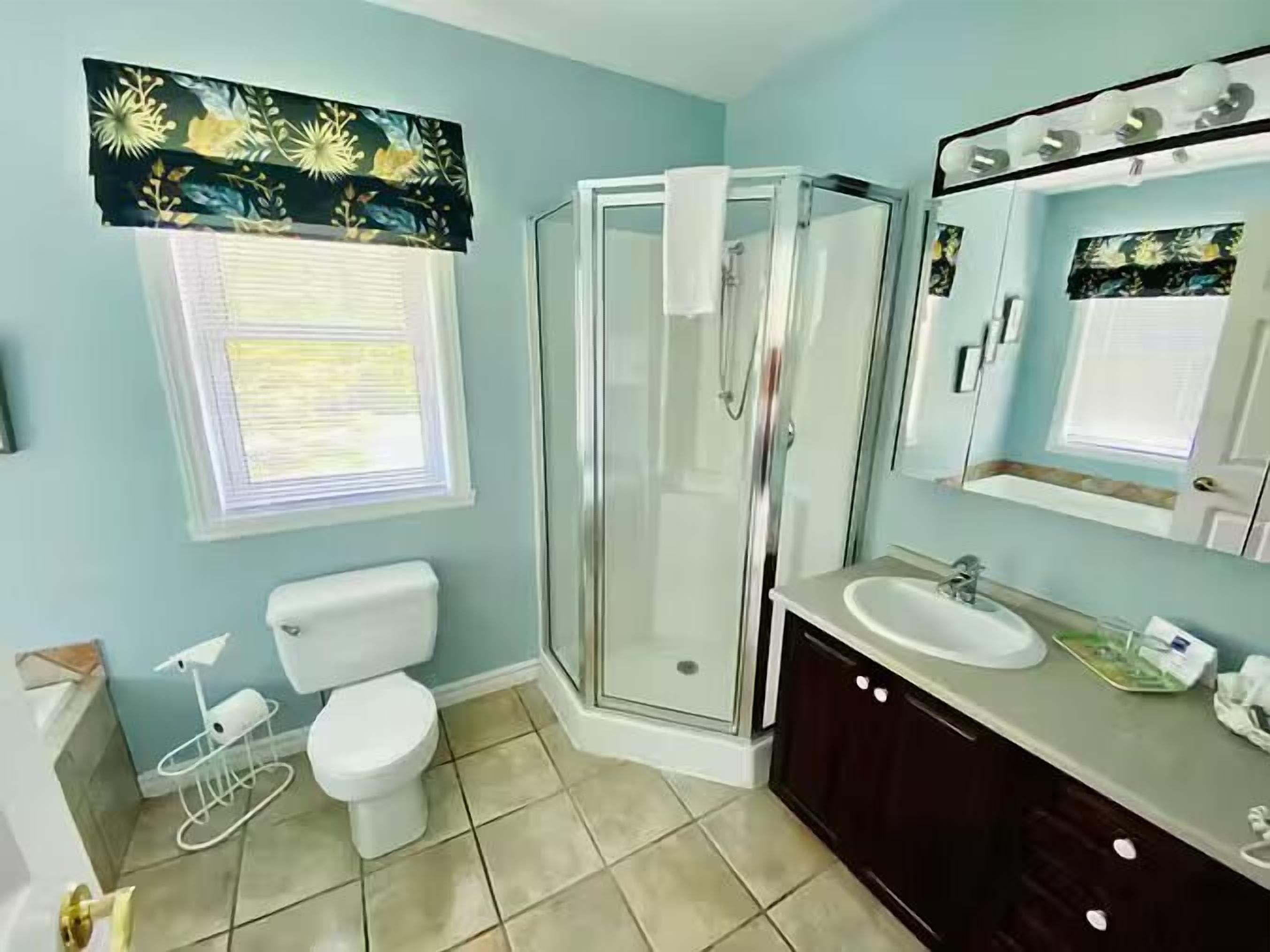 A bathroom with a toilet, shower, sink, and a window with a floral curtain.
