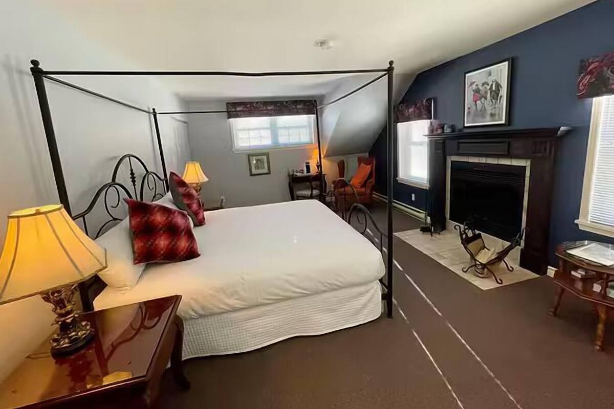 A four-poster bed with white linens and red pillows, a small table with a lamp, a fireplace, and a window with blinds.