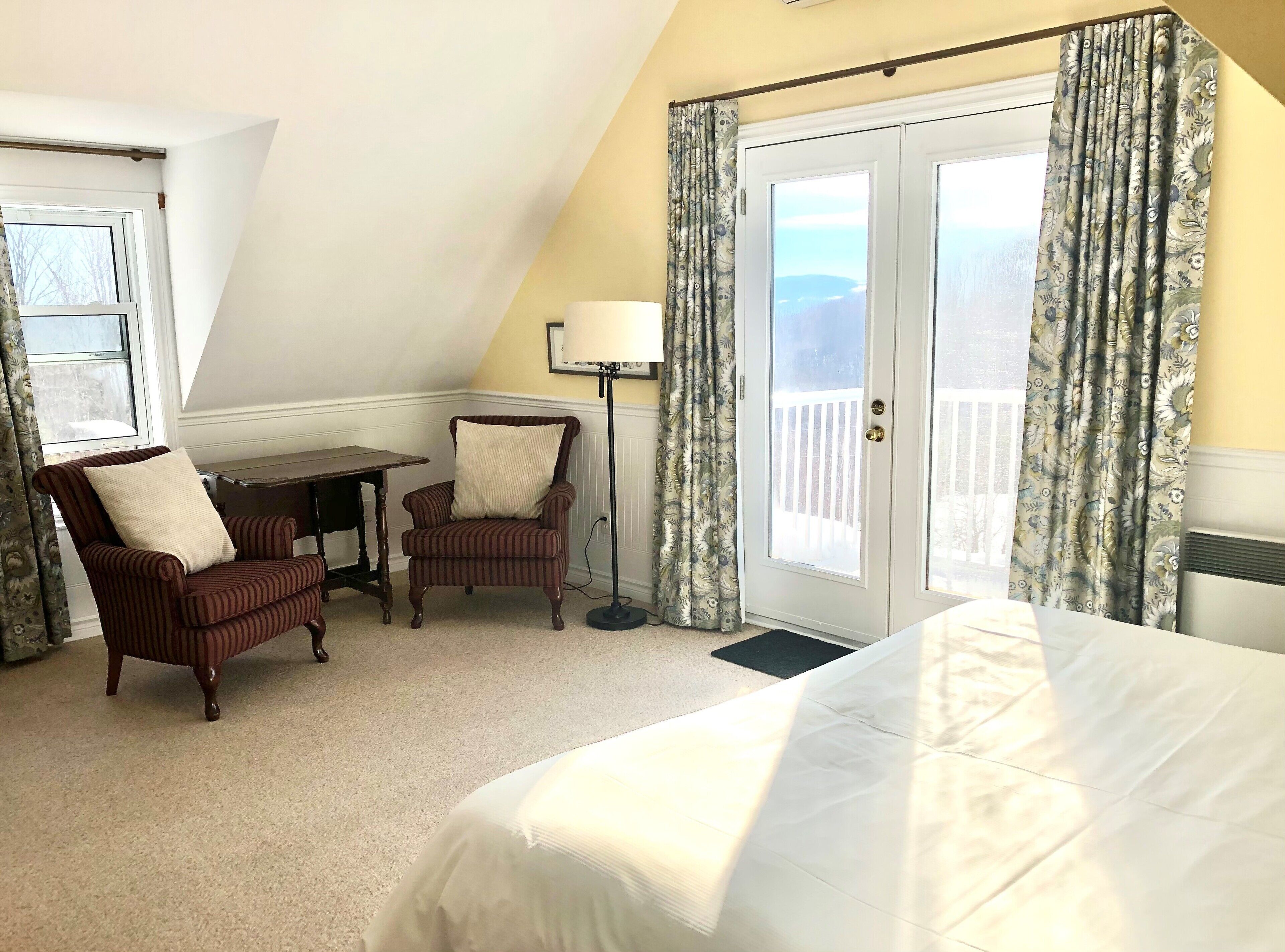 A bedroom with a bed, two chairs, a desk, a floor lamp, and a balcony door with curtains.
