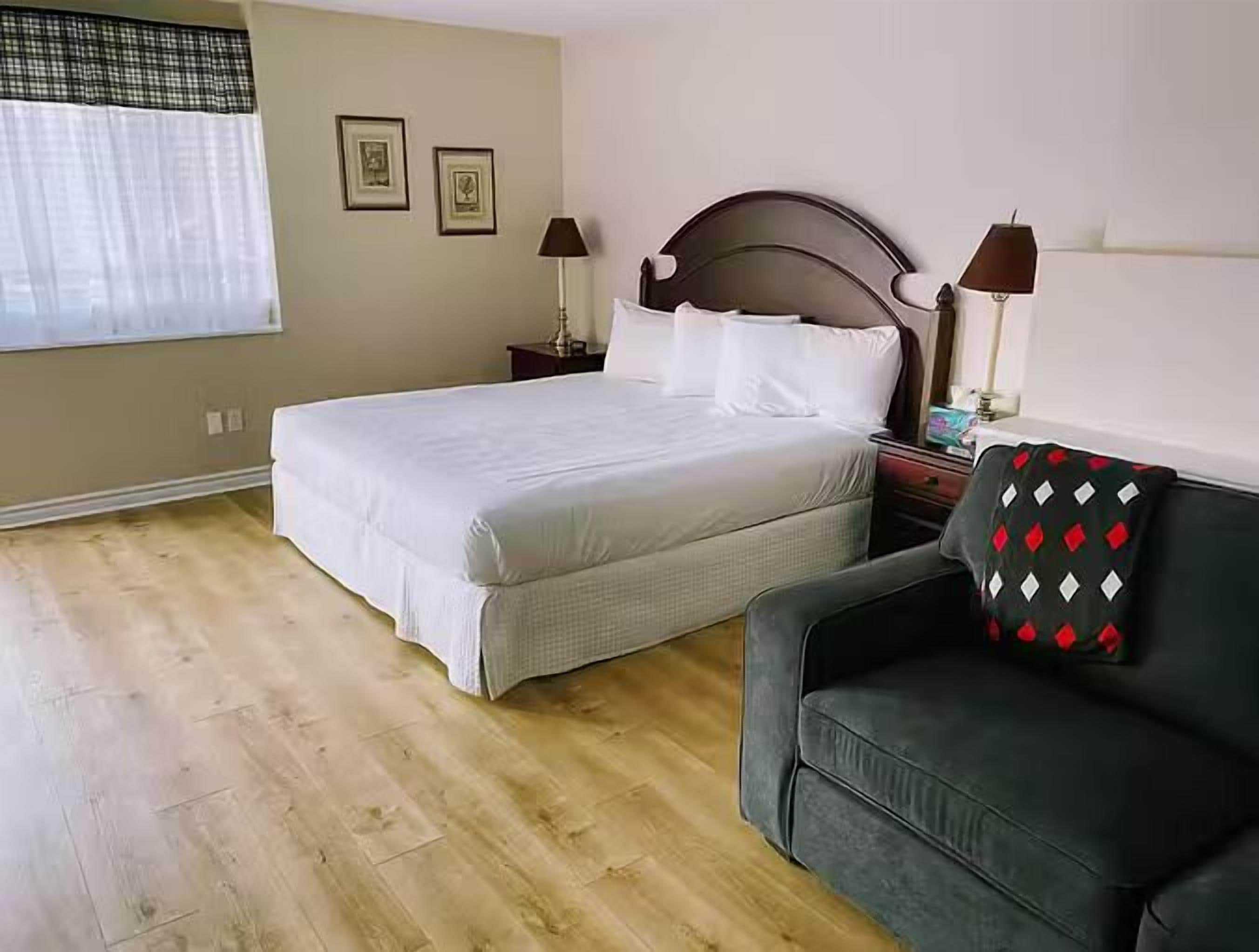 Une chambre à coucher avec un lit, deux tables de chevet avec des lampes, une fenêtre avec des rideaux et un canapé recouvert d’un plaid à motifs.