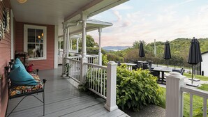 A covered porch with a white railing, a bench with colorful cushions, and outdoor tables and chairs. - Auberge Le Tricorne (Hatley)
