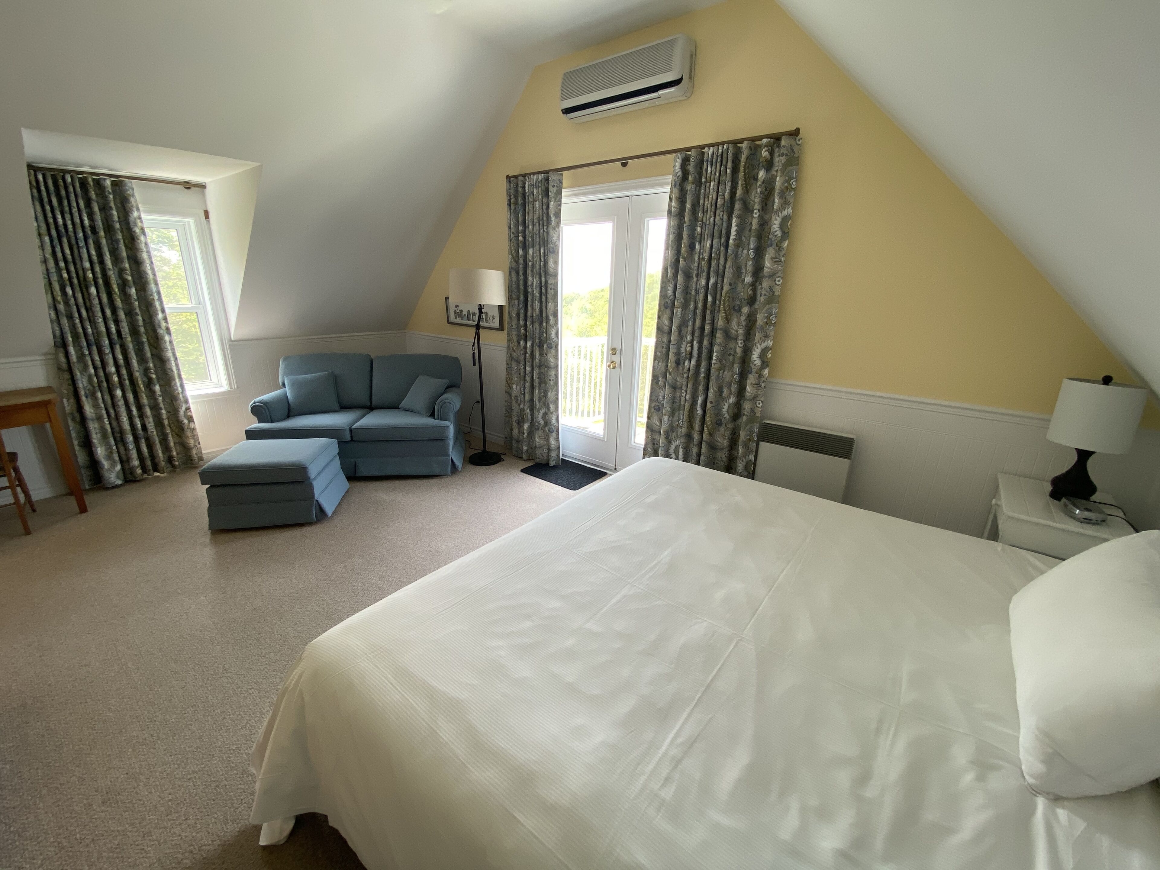 A bedroom with a large bed, a sofa, and a chair. There is a window with curtains, a lamp, and an air conditioning unit on the wall.
