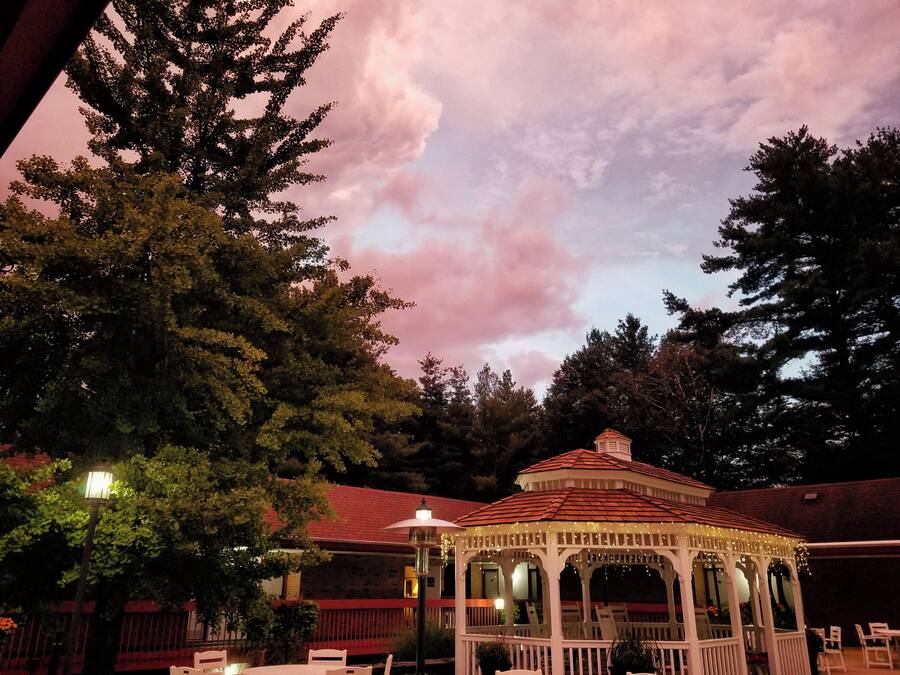 A gazebo with a red roof and white walls, surrounded by trees and a brick building.