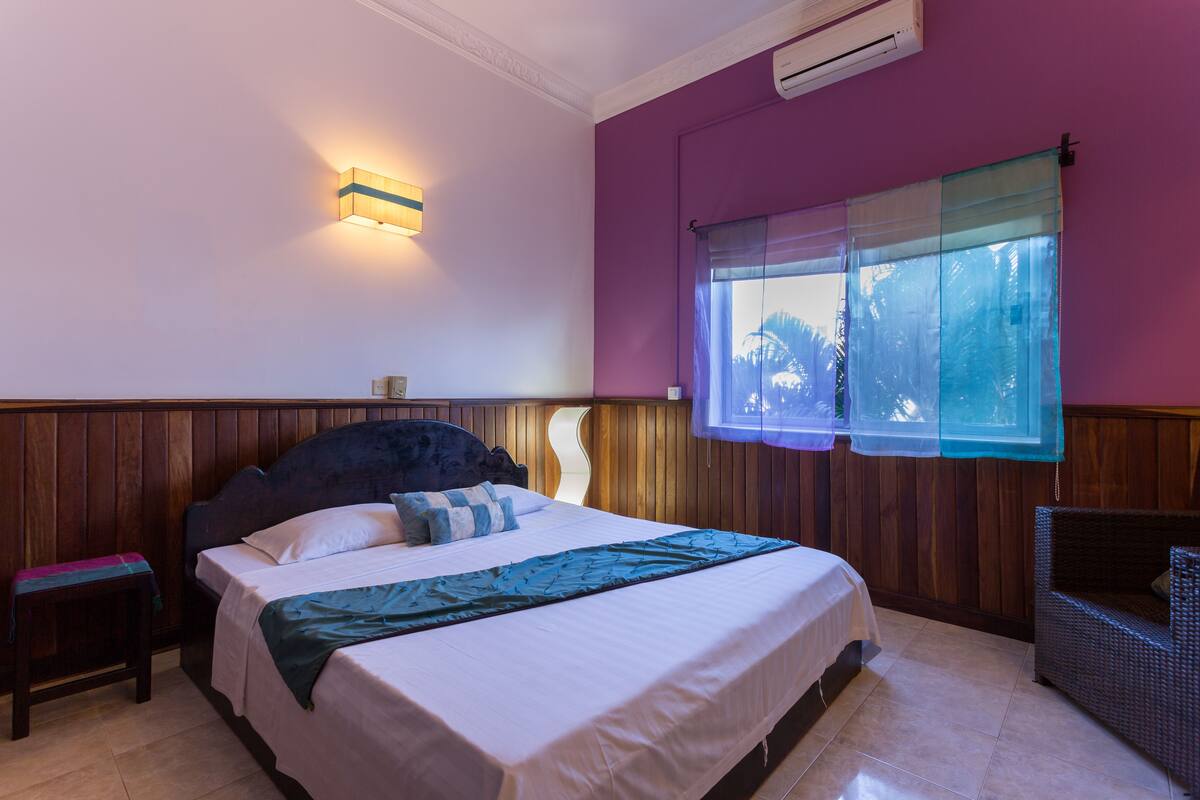 A bedroom with a bed, a nightstand, a window with sheer curtains, and an air conditioning unit on the wall.
