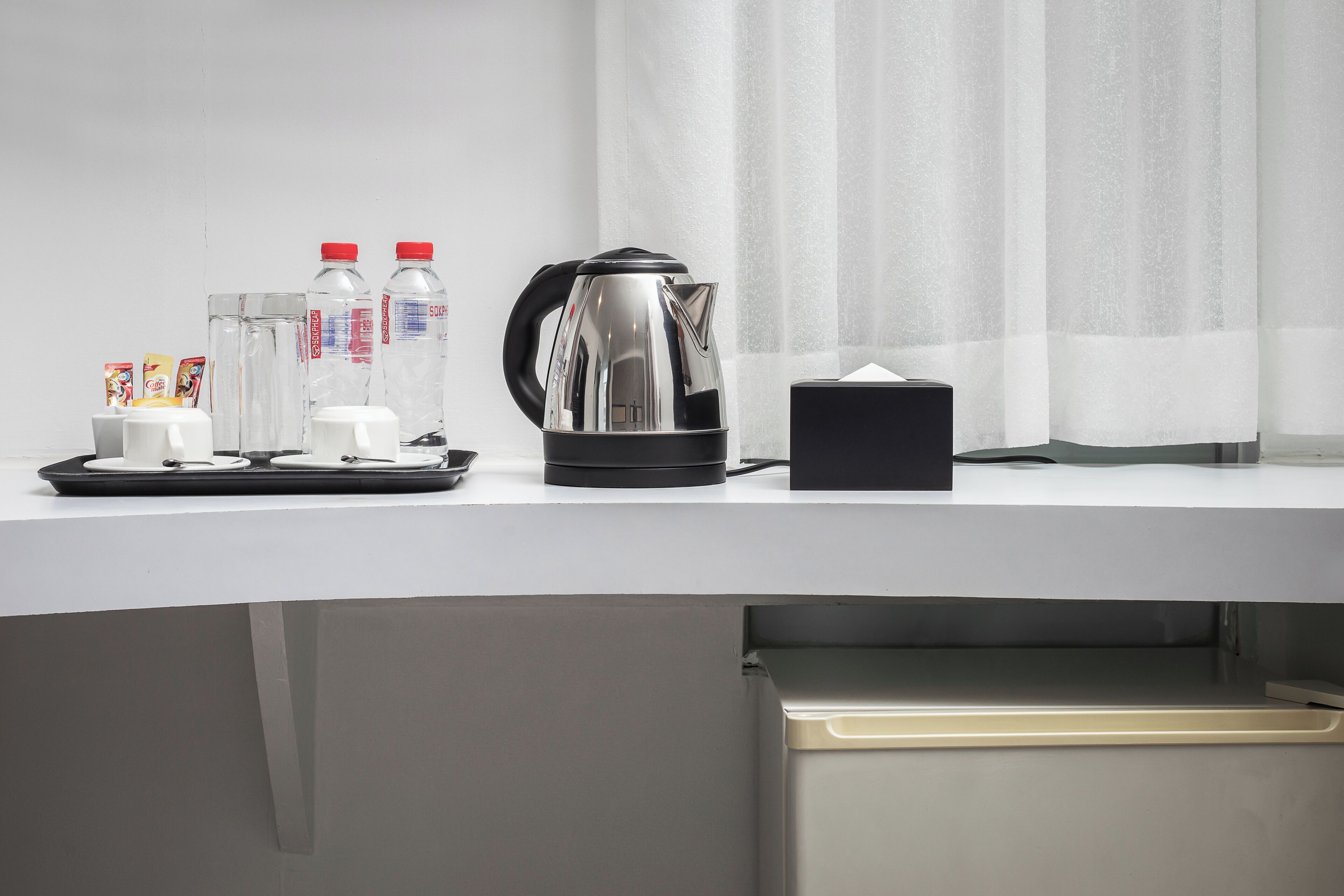Une chambre d’hôtel avec un bureau blanc, une bouilloire, deux bouteilles d’eau, un verre et un plateau avec des tasses et une boîte de mouchoirs.