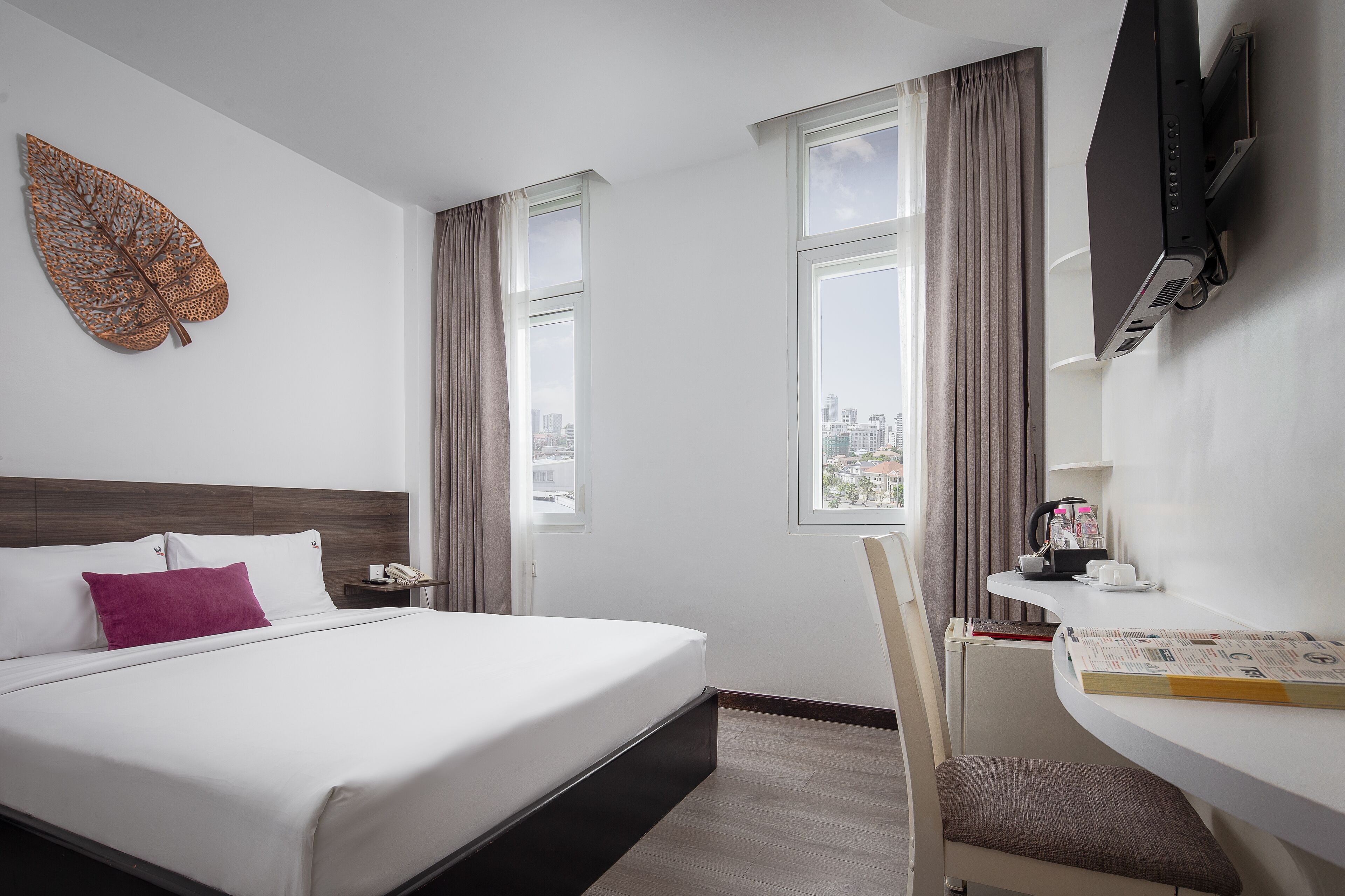 A hotel room with a bed, a desk with a chair, a TV, and a window with curtains.