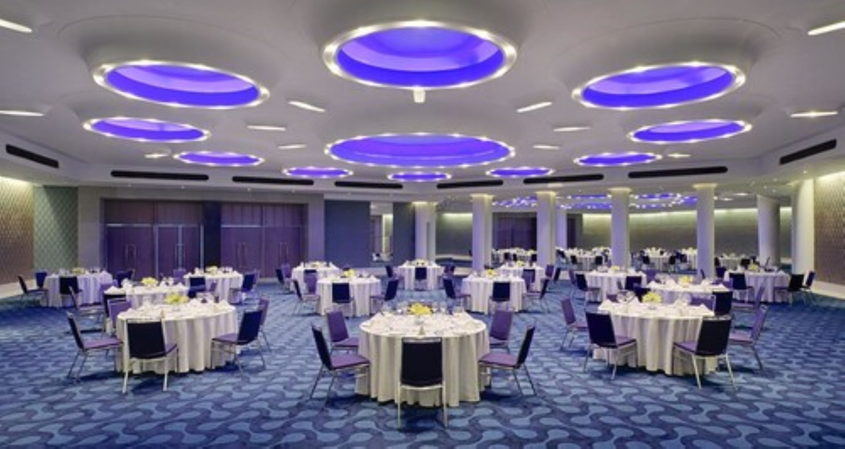 A large banquet hall with round tables and chairs, purple lighting on the ceiling, and a blue carpet.