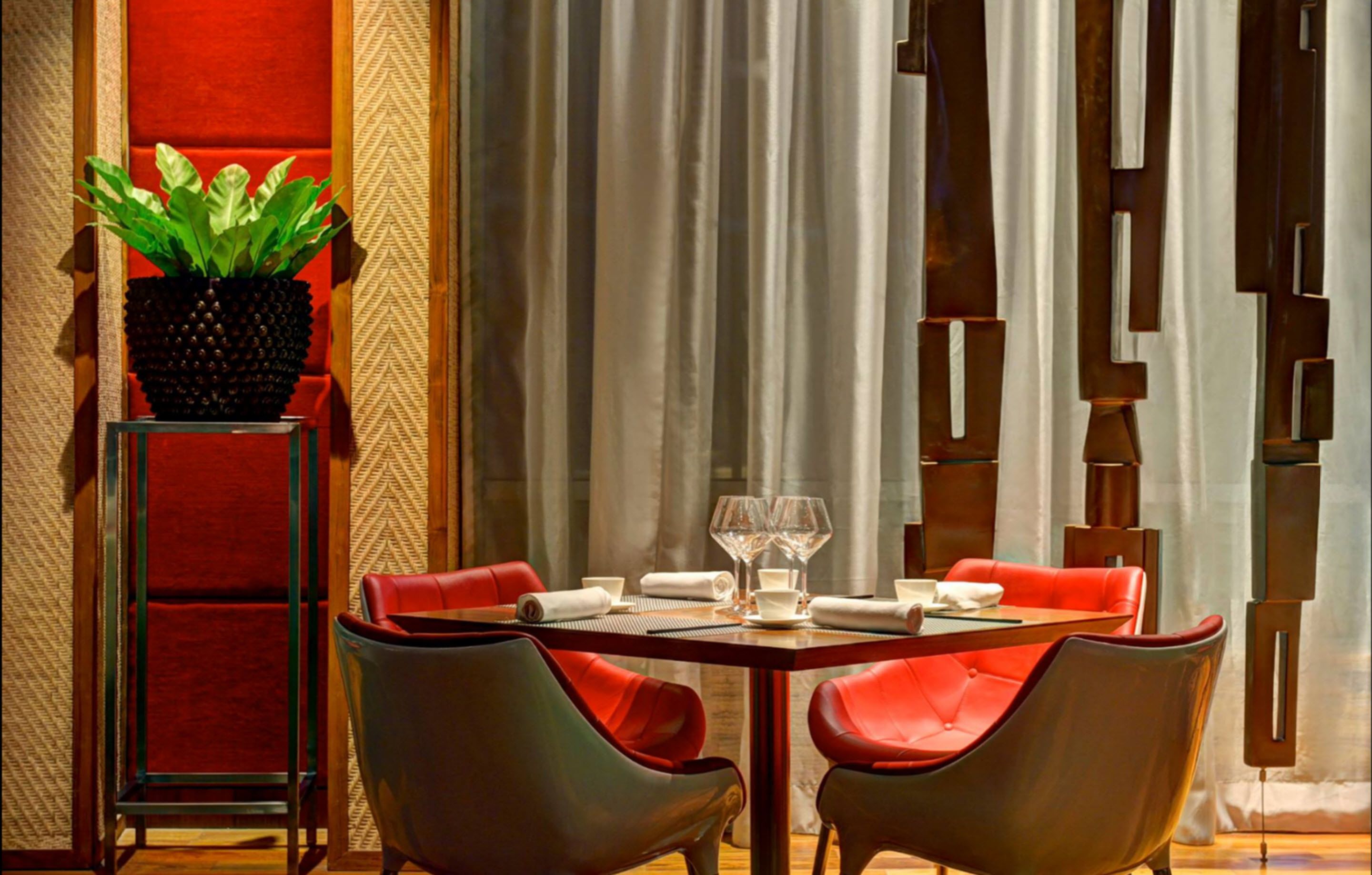 Un comedor con una mesa dispuesta para dos personas, sillas tapizadas en rojo, una planta en maceta y un diseño moderno en la pared.