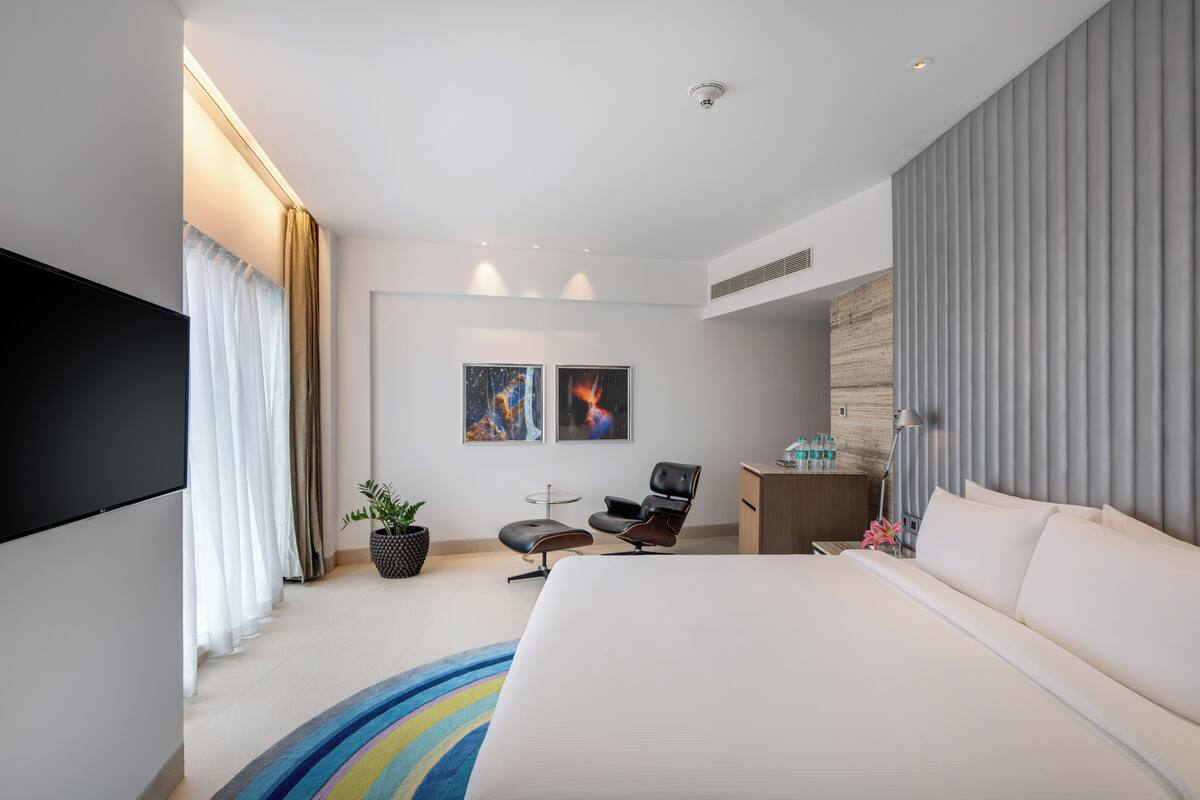 A modern hotel room with a large bed, a desk, a chair, a flat-screen TV, and two framed artworks on the wall.